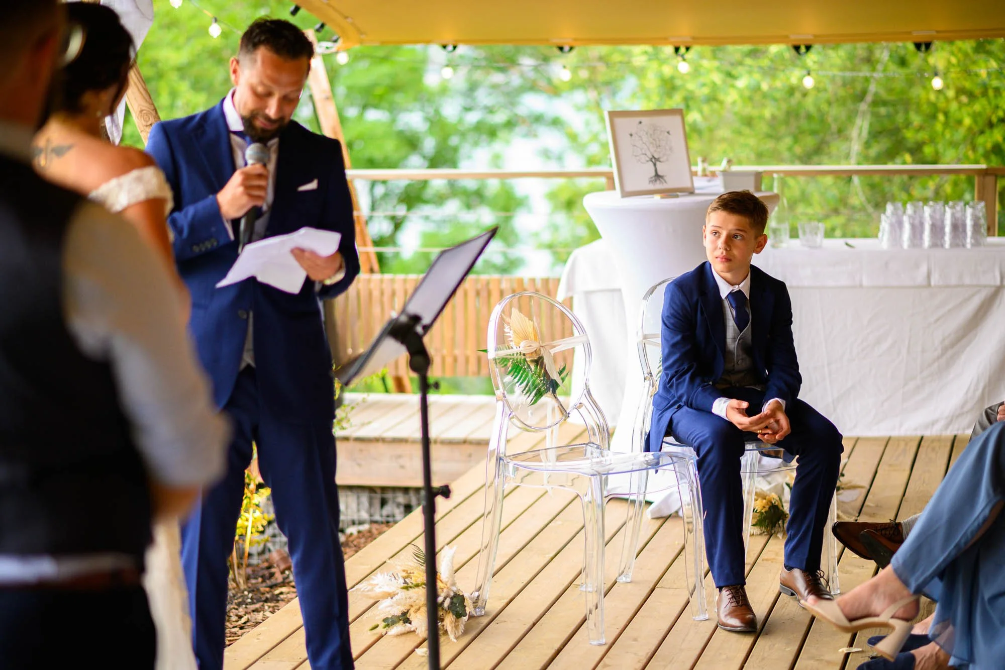Un garçon habillé en costume bleu assis sur une chaise en acrylique lors d'une cérémonie en plein air, avec un homme debout parlant dans un micro. L'événement se déroule sous un abri en bois avec des décorations florales.