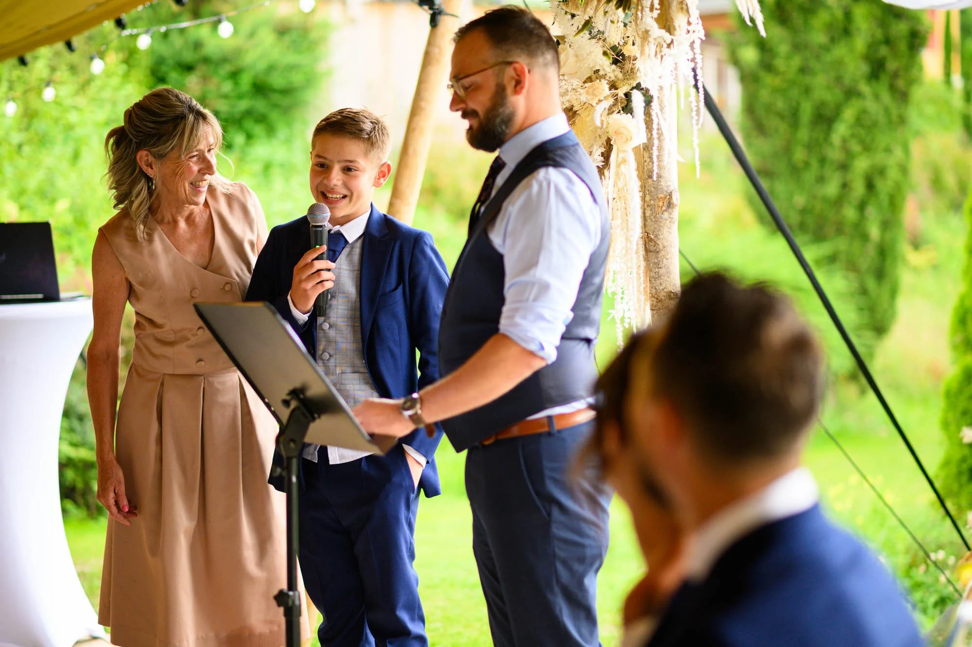 Un garçon en costume bleu parlant dans un micro lors d'une cérémonie en plein air, entouré de personnes souriantes, un homme utilisiant un ordinateur portable, et une femme âgée. Décor de cérémonie avec des fleurs et une forêt en arrière-plan.