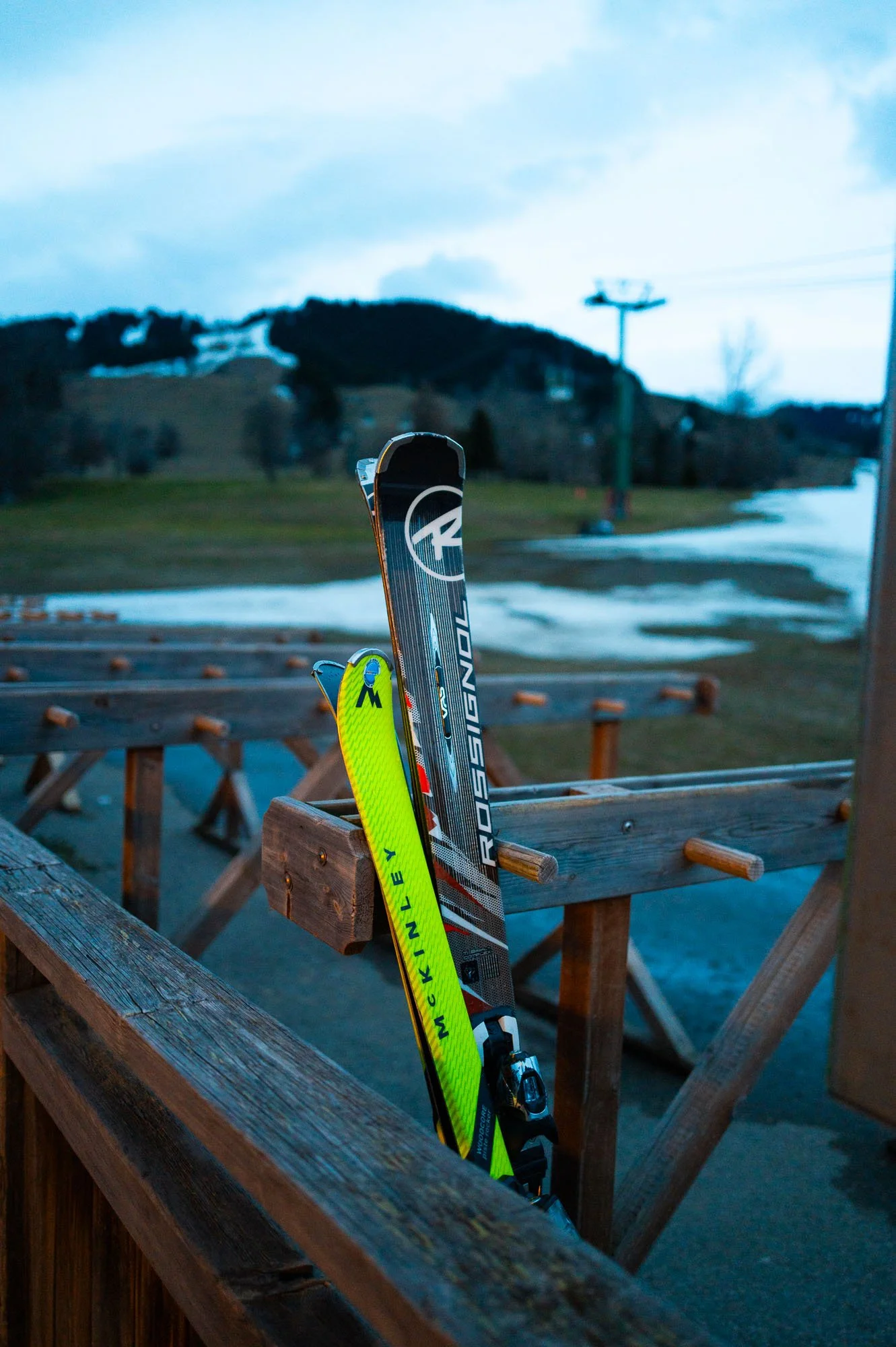 Deux skis posés contre une barrière en bois à une station de ski. Le paysage en arrière-plan montre des montagnes, des pistes de ski, et un ciel nuageux, avec quelques traces de neige au sol.