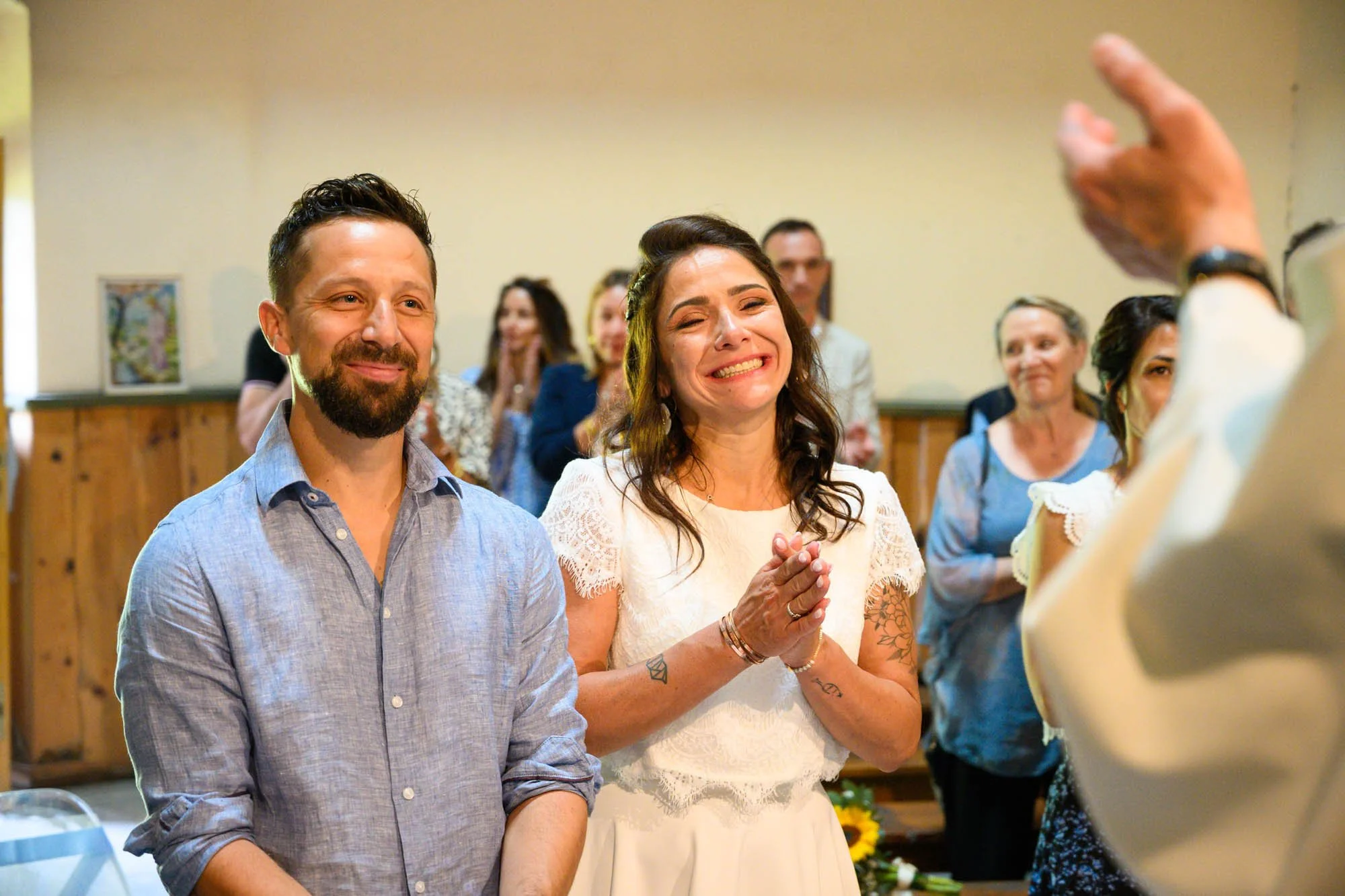 Un couple souriant à un mariage, entouré de convives en admiration.