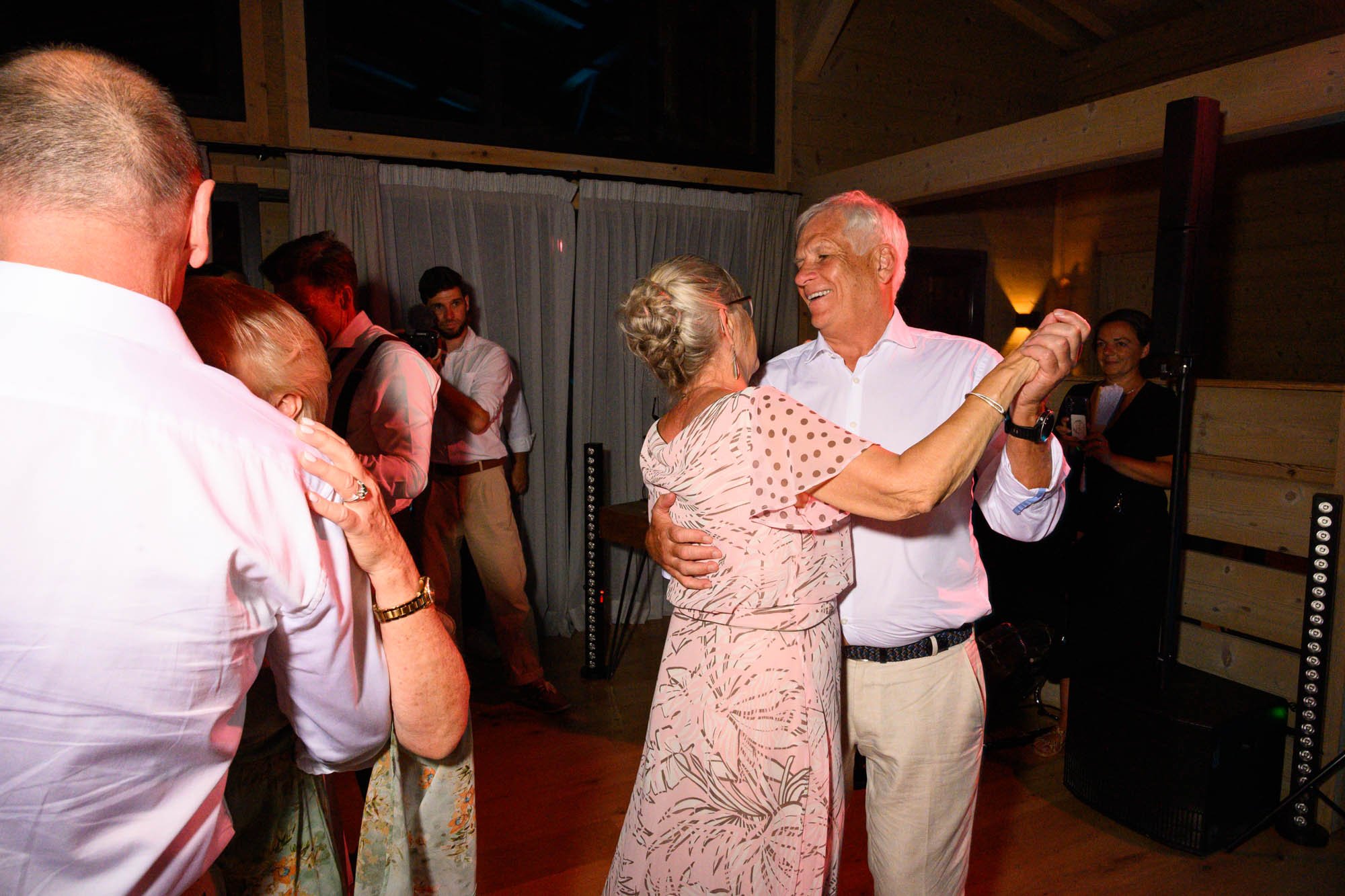 Un couple danse lors d'une fête, souriant et portant des vêtements décontractés, dans une salle en bois, avec d'autres personnes en arrière-plan.