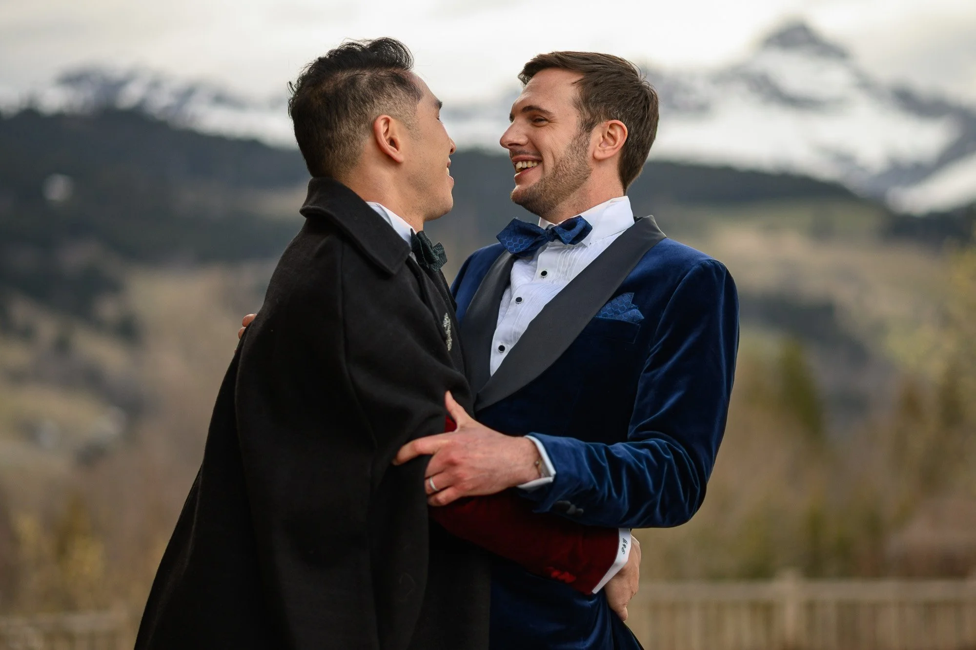 Deux hommes habillés élégamment, se tenant la main et souriant, dans un décor naturel avec des montagnes enneigées en arrière-plan.
