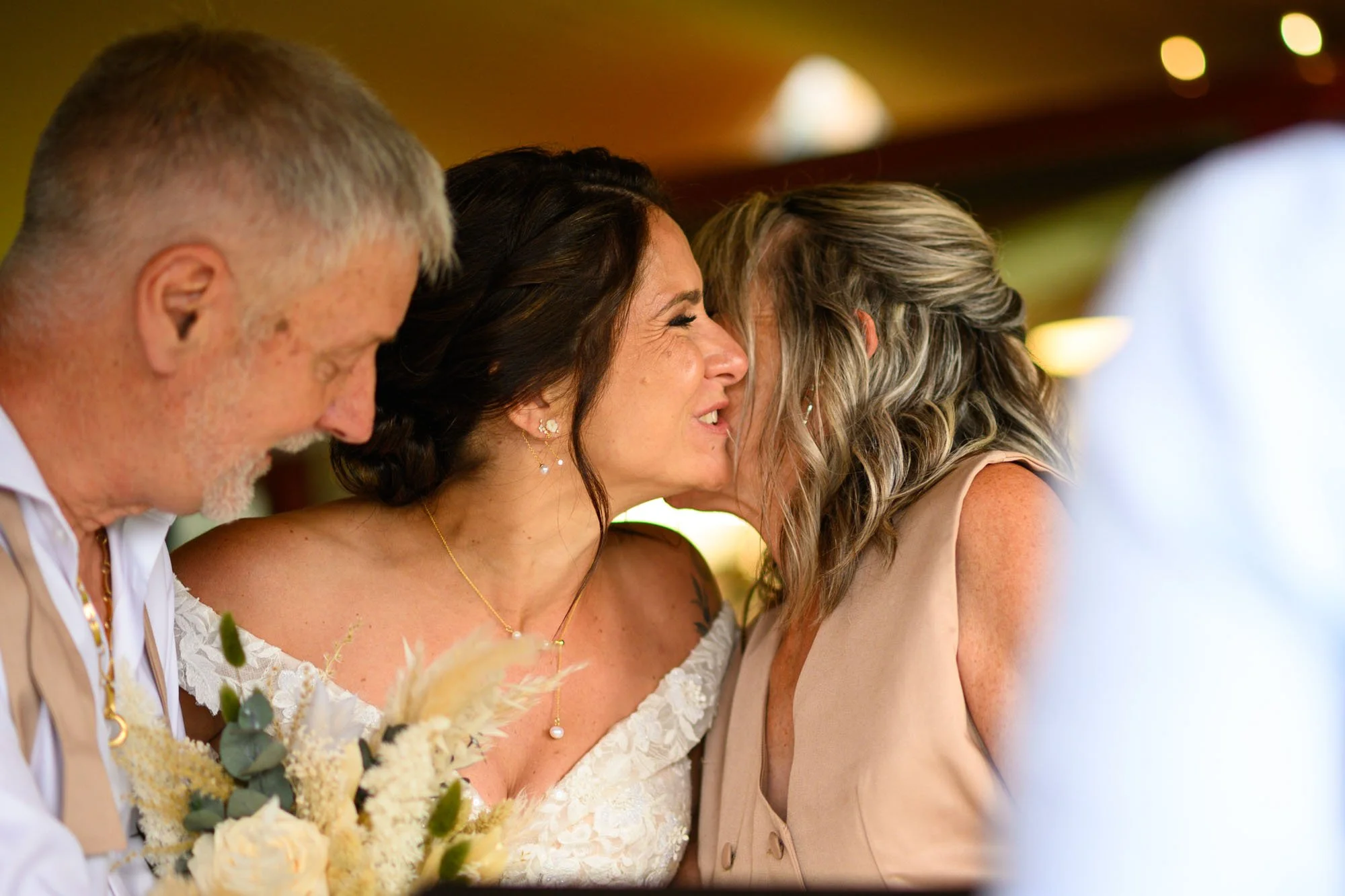 Deux femmes et un homme à une fête, la femme au centre a une robe blanche et sourit en étant biseautée avec une autre femme portant une tenue beige, l'homme à gauche a une barbe blanche et porte une chemise blanche.