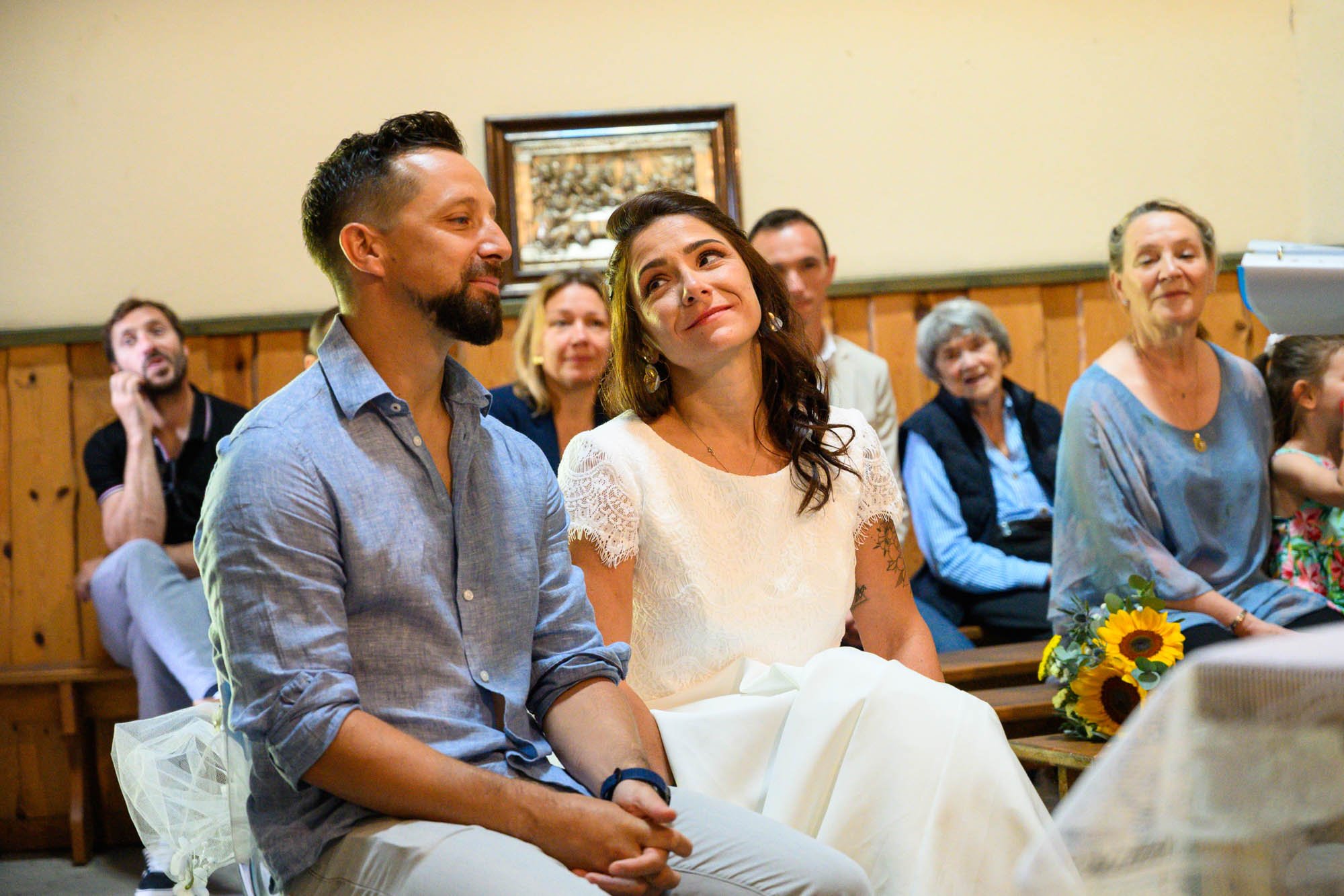Un couple assis lors d'une cérémonie, entouré de plusieurs personnes assises en arrière-plan, dans une pièce avec un mur en bois.
