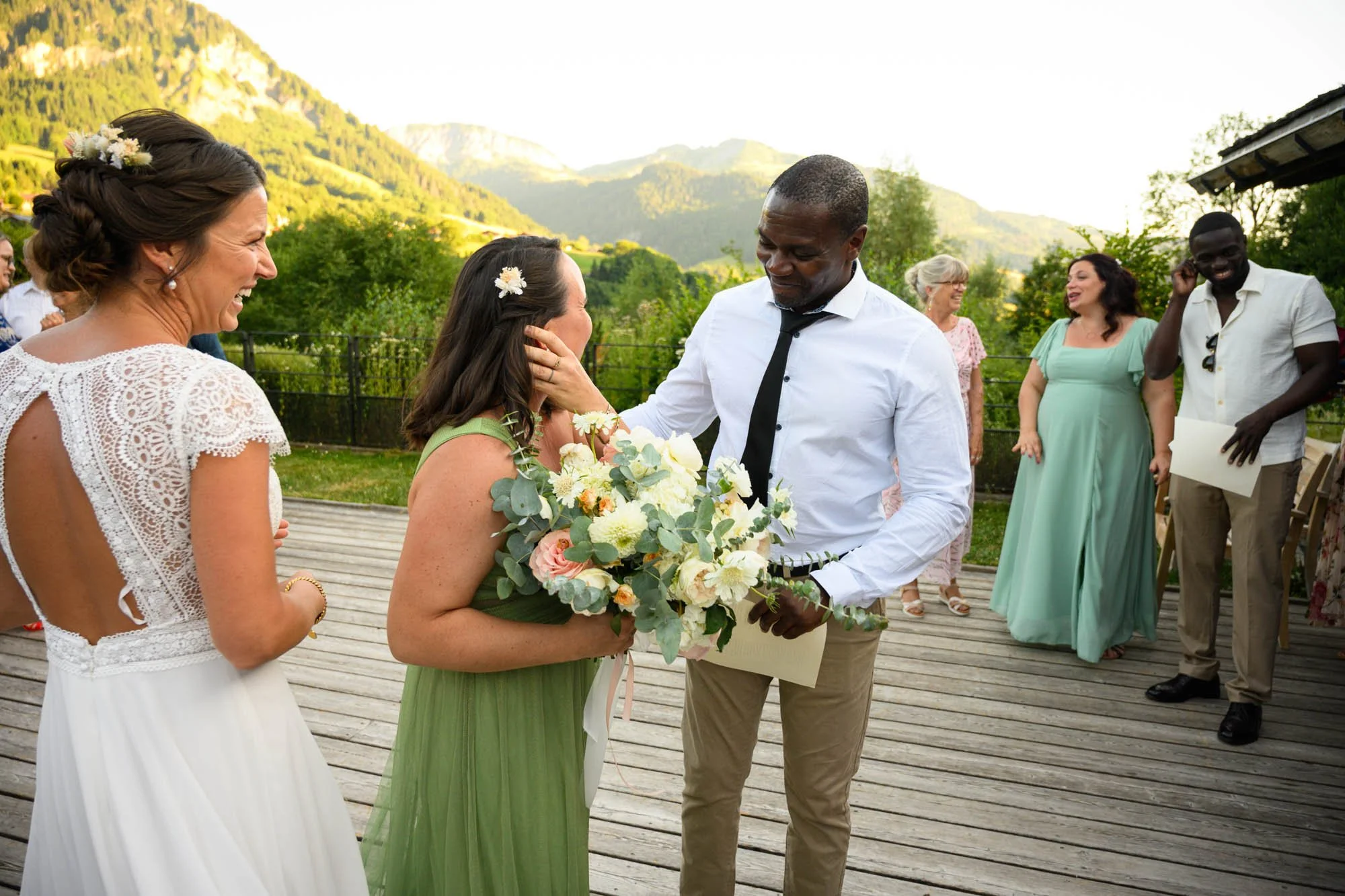 Un homme offre un bouquet de fleurs à une femme lors d'une cérémonie en plein air, avec d'autres personnes en arrière-plan, dans un cadre naturel avec des montagnes en toile de fond.