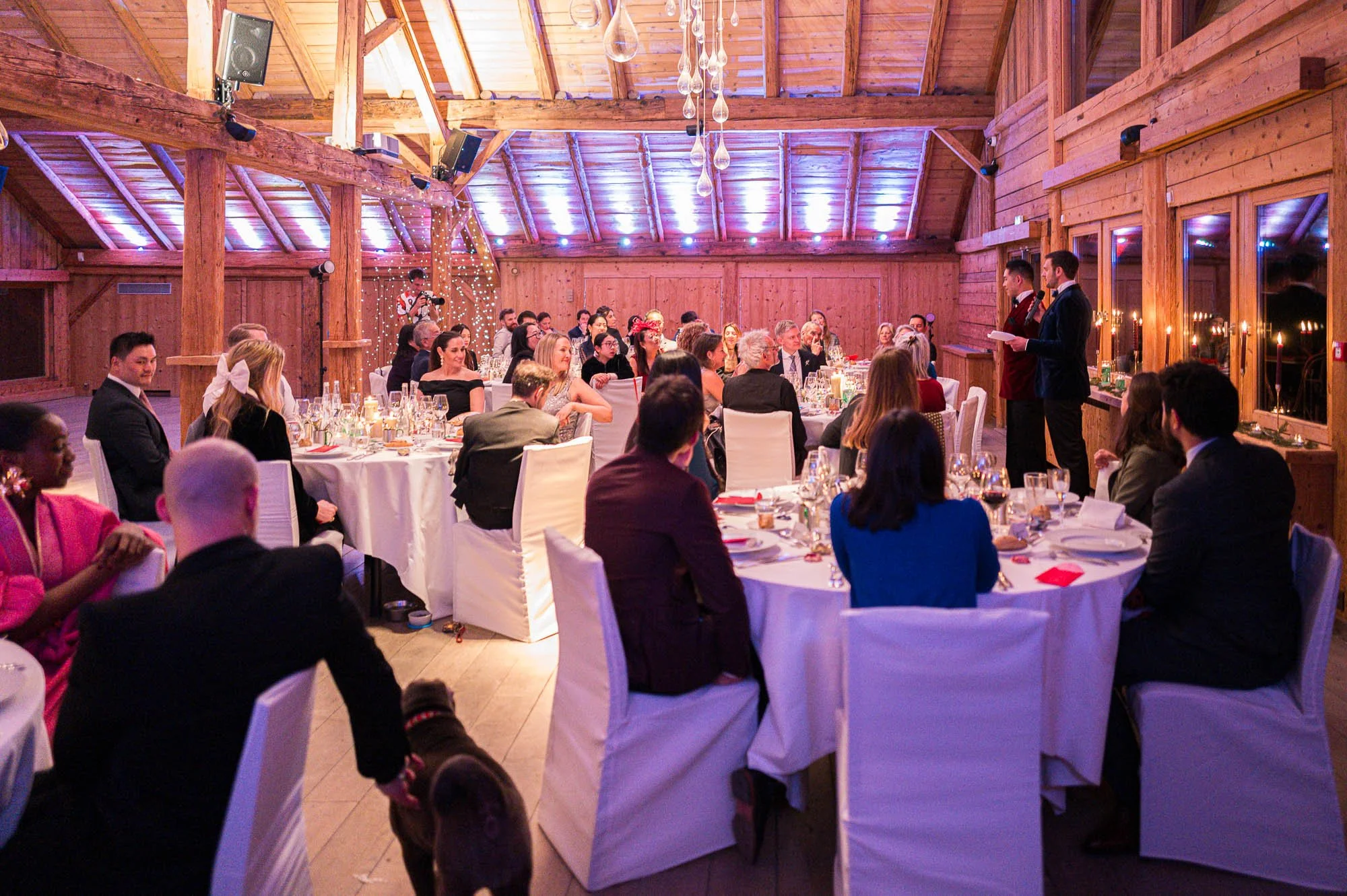 Une salle de réception en bois avec un plafond en pente, décorée pour un événement, où un groupe de personnes assises autour de tables élégantes écoute deux hommes debout en train de faire un discours.