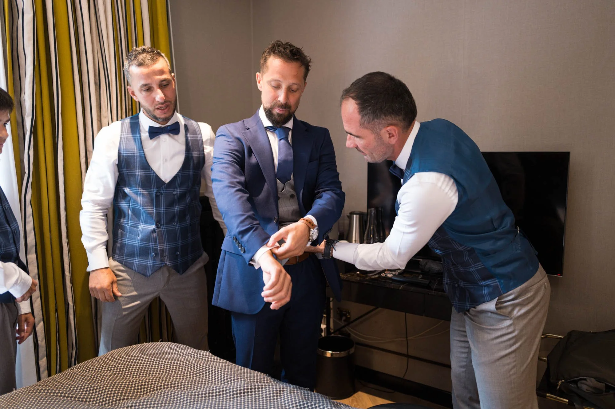 Groom adjusting his cufflinks while a man helps him, with two other men watching, all dressed in suits, in a room with curtains and a television in the background.
