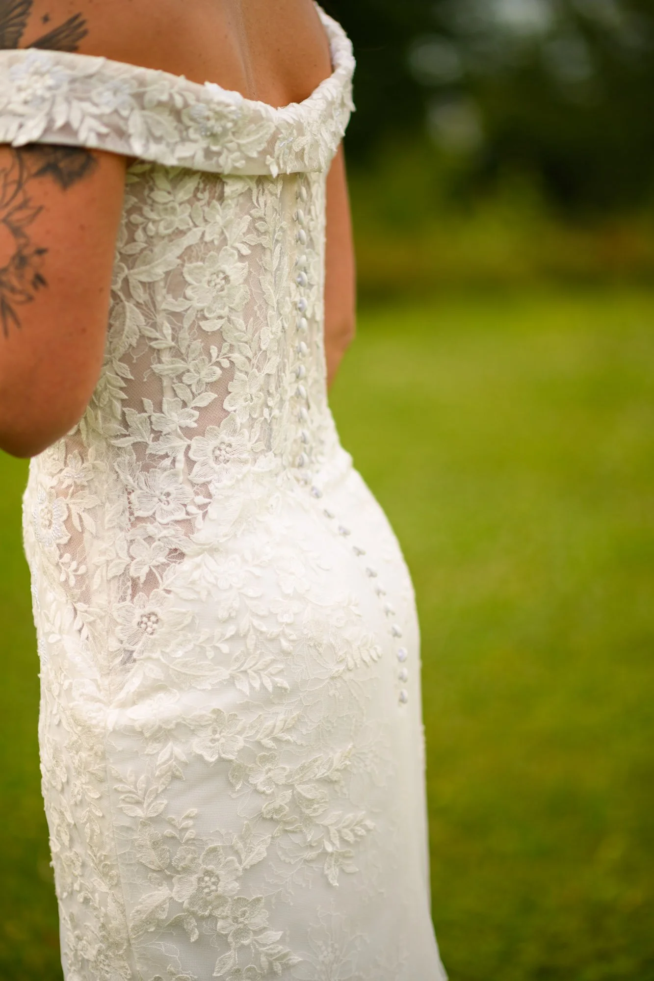 Détail d'une robe de mariage en dentelle blanche avec des boutons en ligne sur le dos, porté par une personne dont le bras, avec un tatouage, est visible.