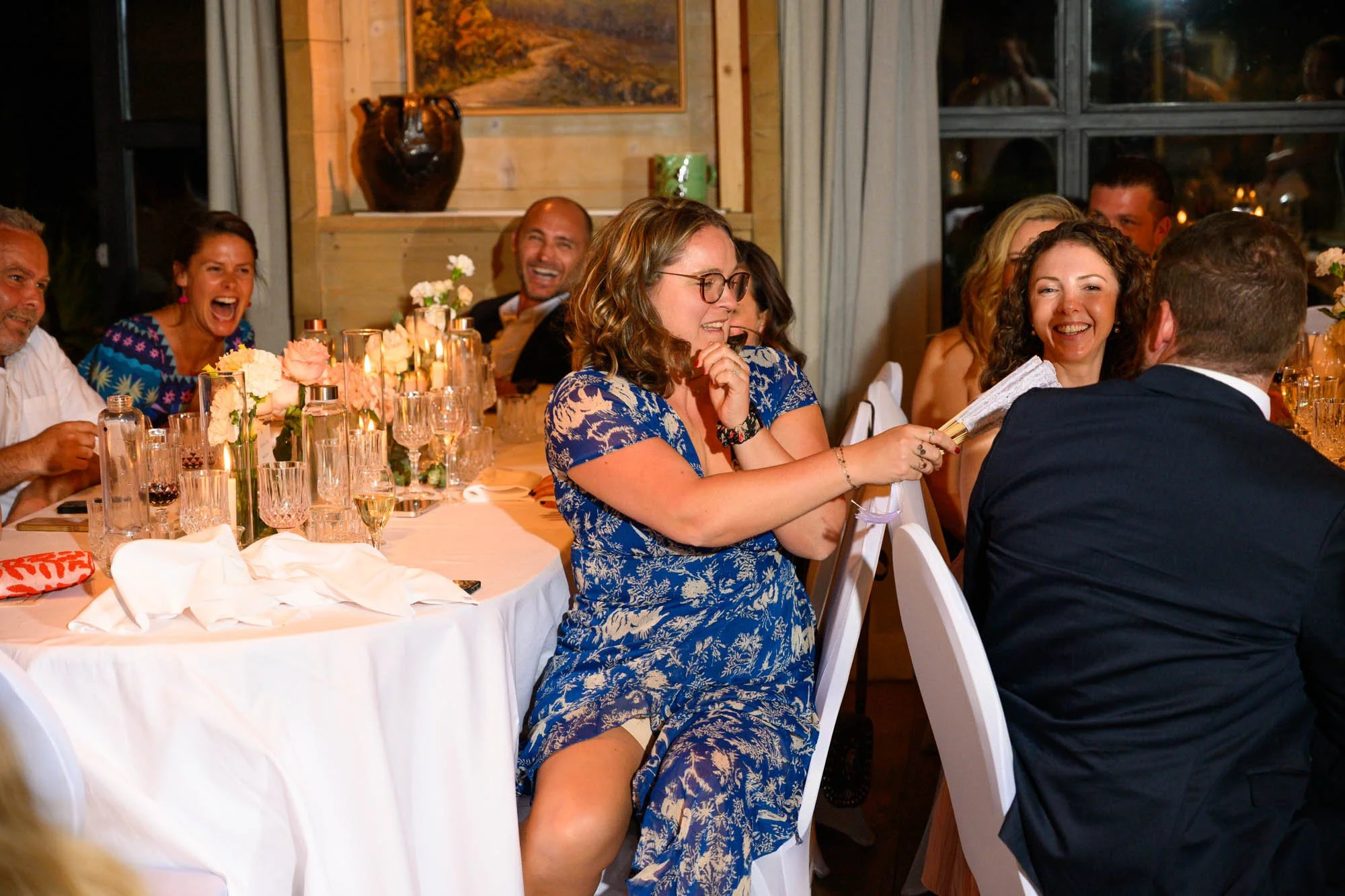 Un groupe de personnes assises à une table lors d'un repas, certains rient, dans une ambiance chaleureuse et festive.