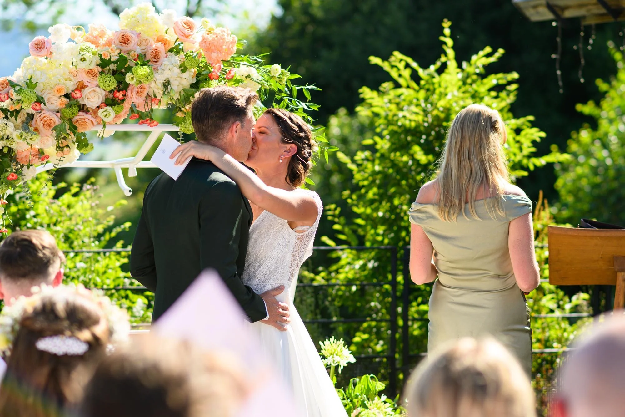 Un couple s'embrasse lors de leur mariage en plein air, entouré de fleurs et de verdure, avec des invités en arrière-plan.
