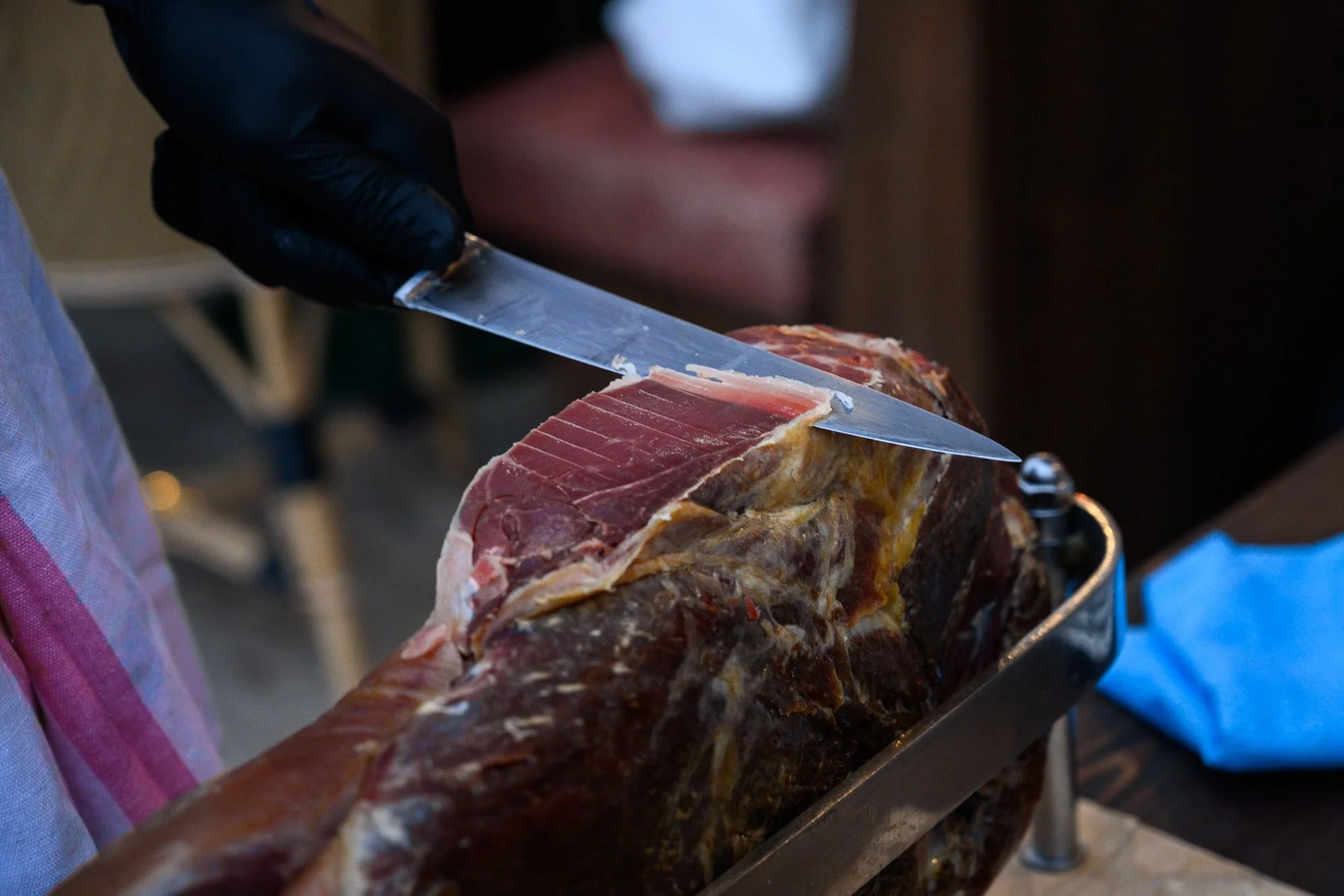 Une personne portant un gant noir coupe du jambon cru avec un couteau dans une charcuterie.