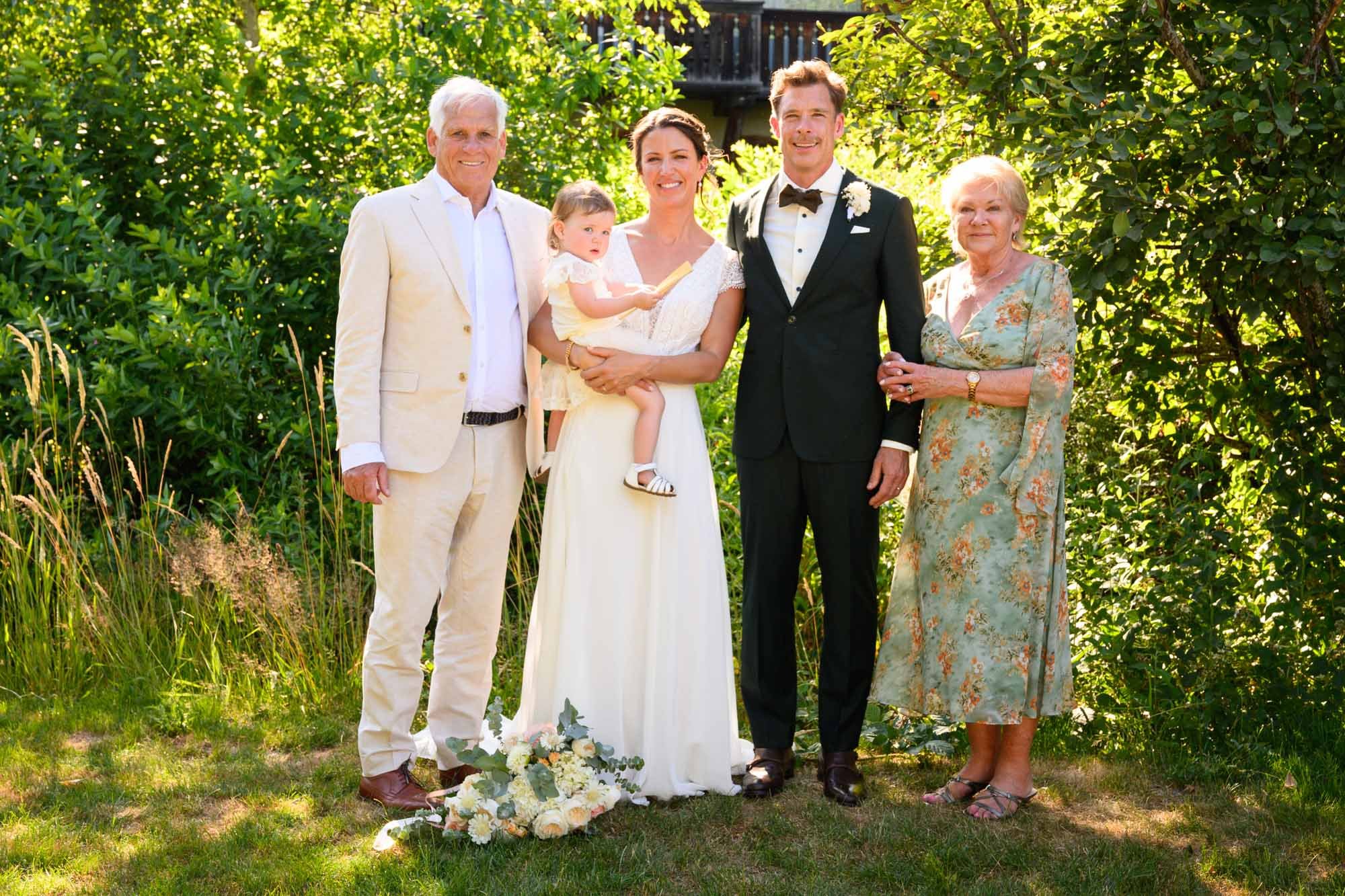 Groupe de six personnes à un mariage en extérieur, avec un grand bouquet blanc sur l'herbe devant eux, fond de feuillage vert.