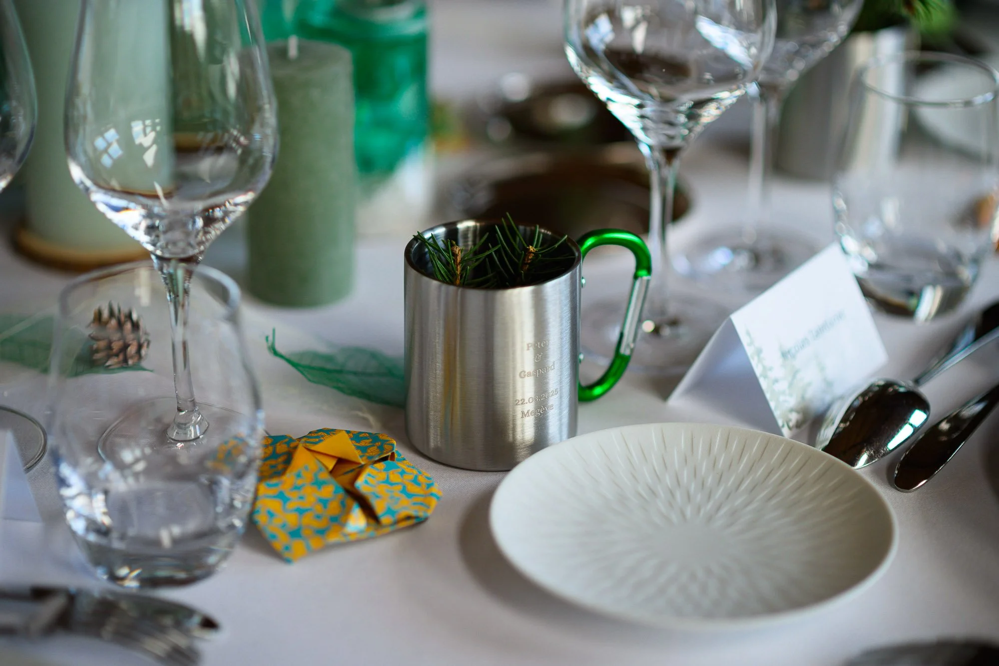 Un centre de table avec un mug en acier inoxydable contenant des branches de pin, entouré de verres à vin, d'assiettes et de couverts, pour un repas ou une réception.