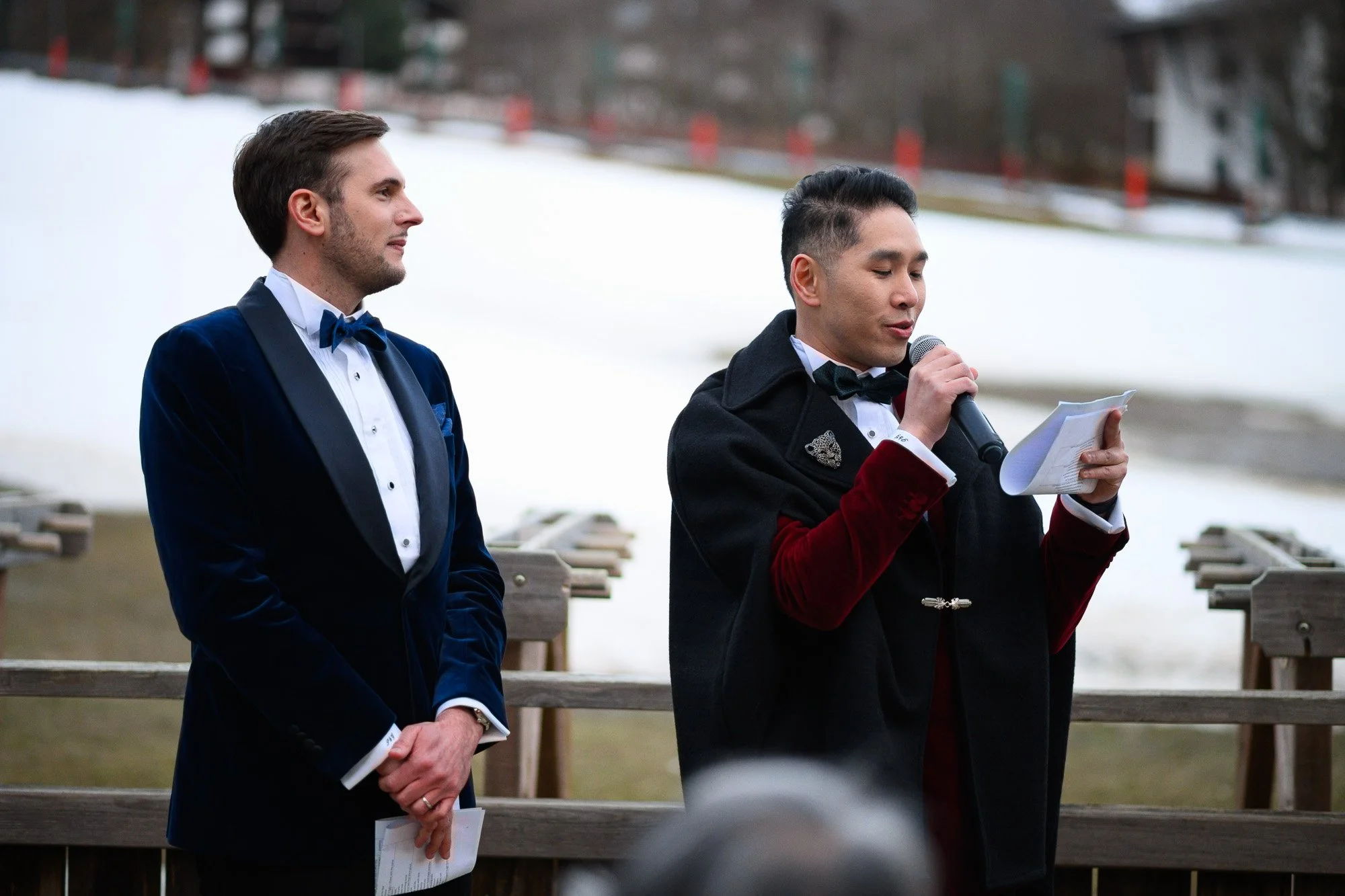 Deux hommes en costumes formels, l'un avec un costume bleu et une cravate bleu, l'autre avec un costume noir et une cravate noire, se tenant à une cérémonie en plein air devant un paysage enneigé, l'homme à gauche regarde l'autre qui lit à haute voix