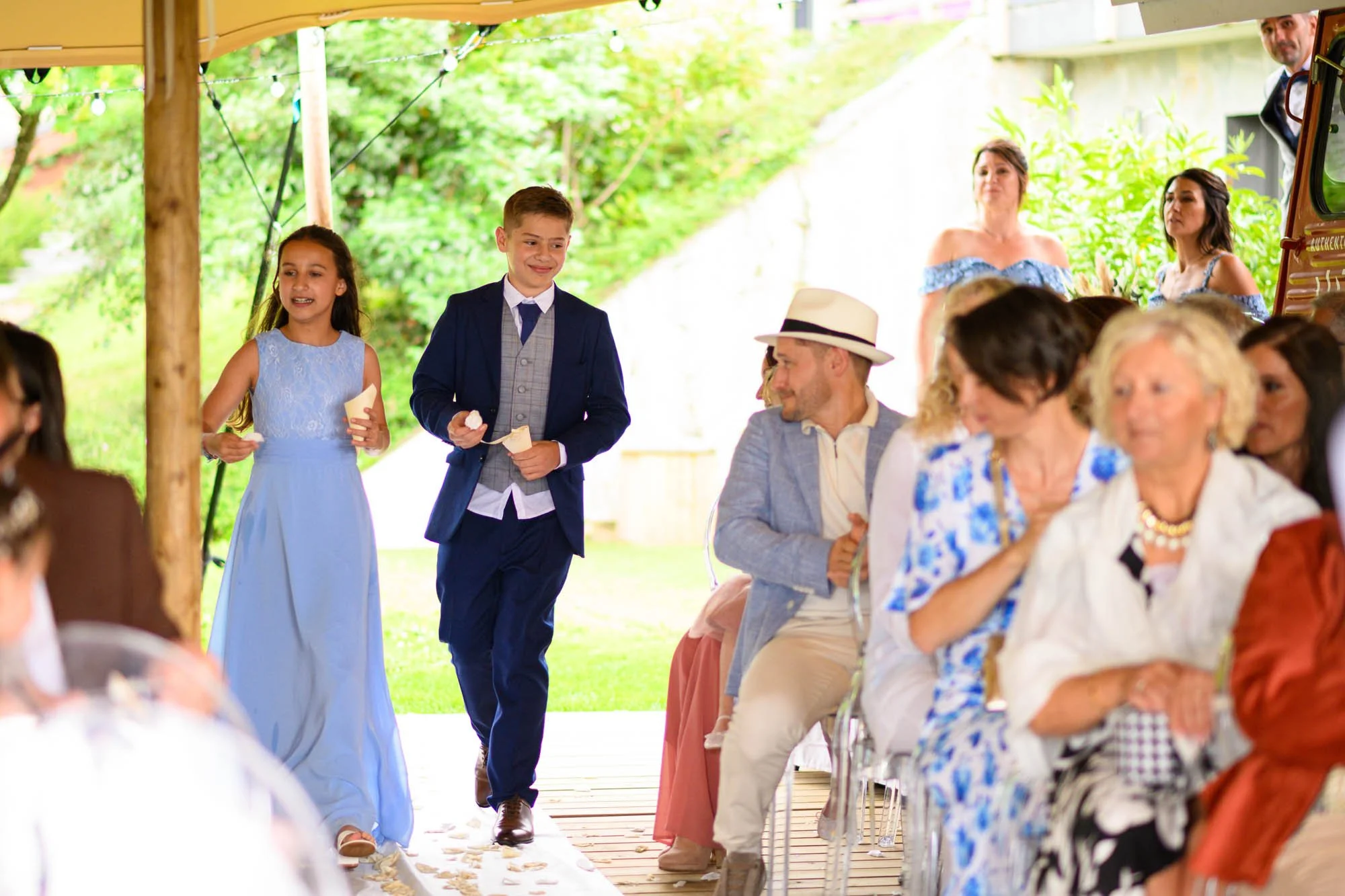 Des enfants en costume marchent dans un couloir lors d'une cérémonie de mariage, entourés de personnes assises et souriantes, sous une tente décorée.