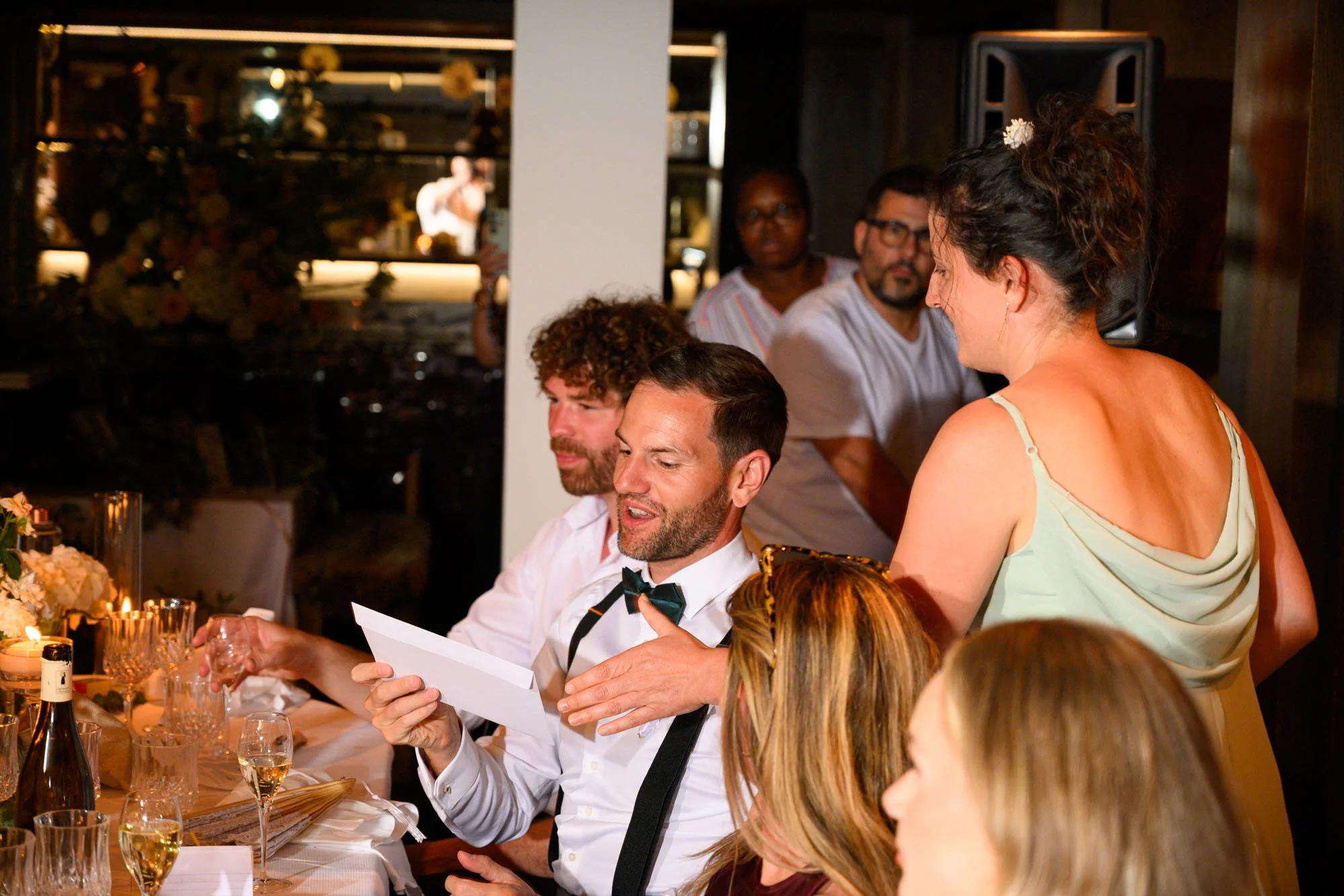 Un homme en smoking avec un nœud papillon donne un discours lors d'un repas, entouré d'autres invités, dans un restaurant ou une salle de réception