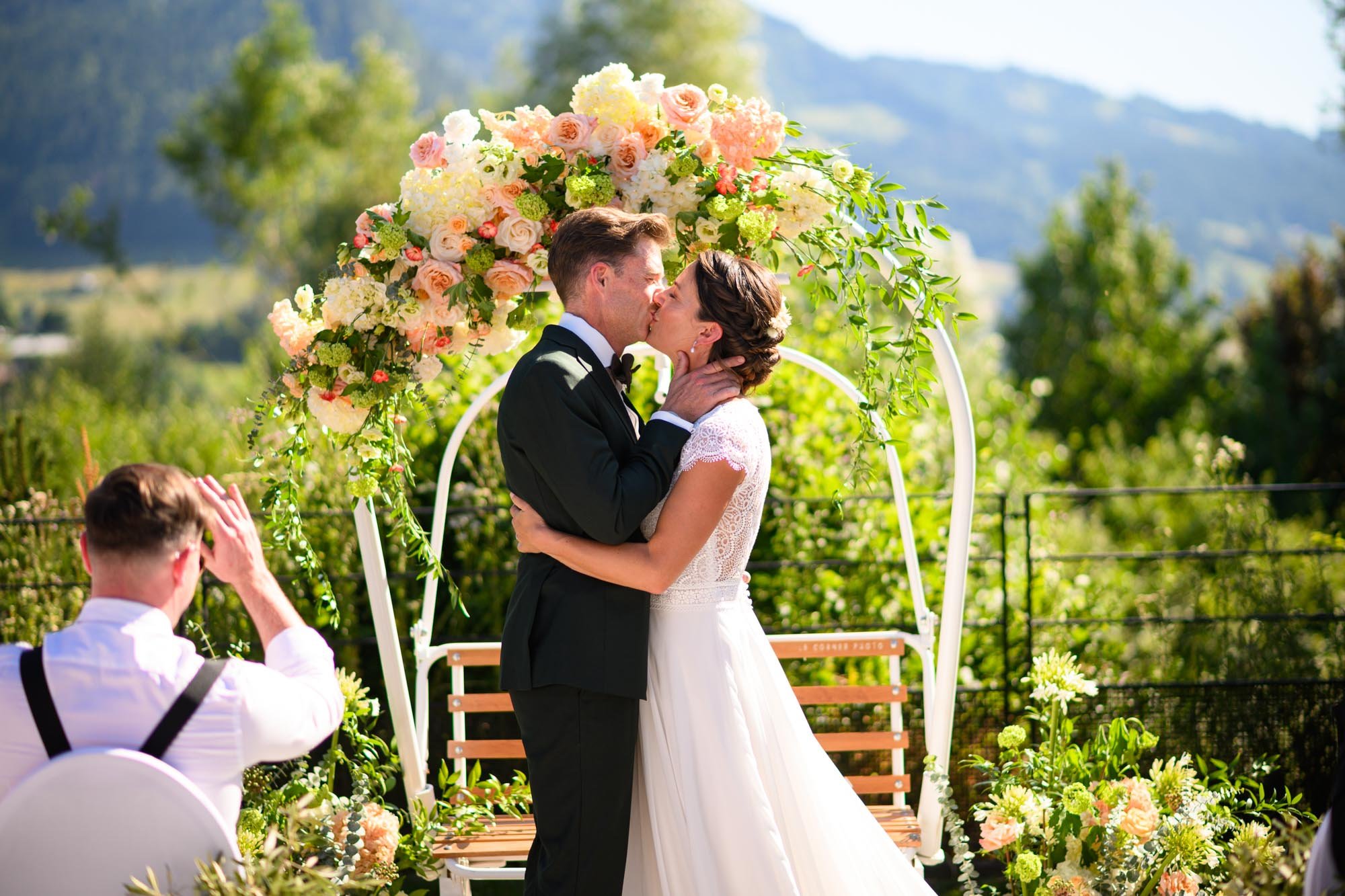 Un couple de mariés s'embrassant lors de leur mariage en plein air, avec un décor floral et un cadre naturel verdoyant.