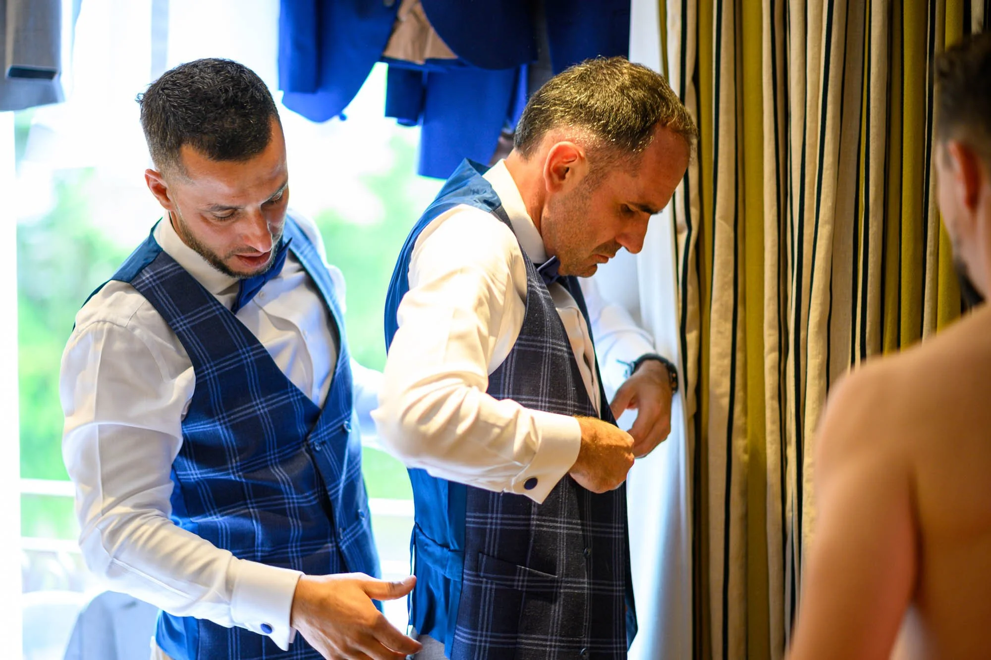 Deux hommes en costume vérifient la tenue de leur compagnon à l'aide d'une conversation dans une pièce lumineuse avec des rideaux jaunes suspendus.