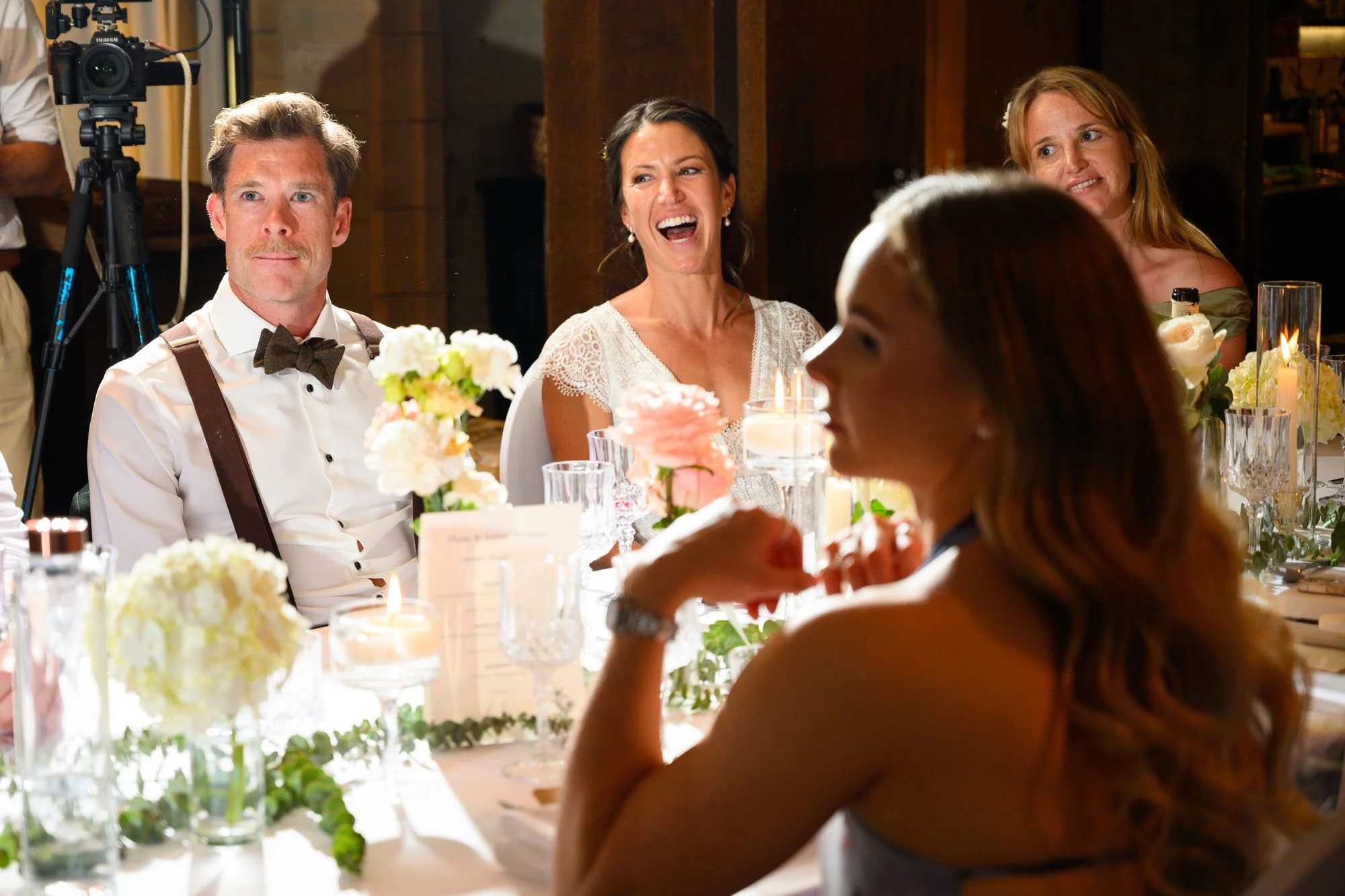 Groupe de personnes assises à une table lors d'un mariage ou d'une célébration, avec des fleurs et des bougies, certaines personnes souriant ou riant.