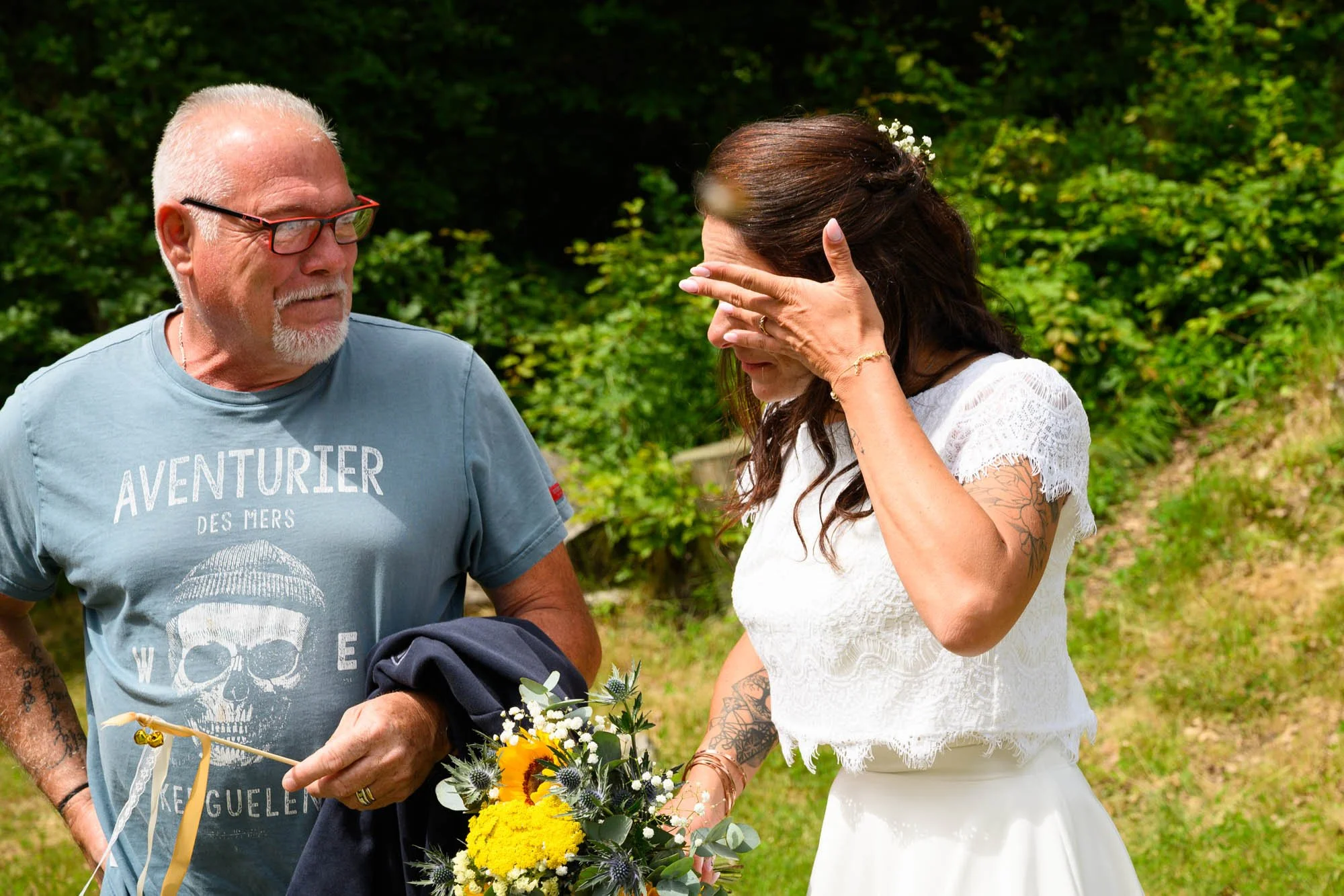 Une femme en robe blanche avec des tatouages et des fleurs dans les cheveux, pleurant en tenant un bouquet, tandis qu'un homme plus âgé lui tend quelque chose lors d'une cérémonie en plein air.