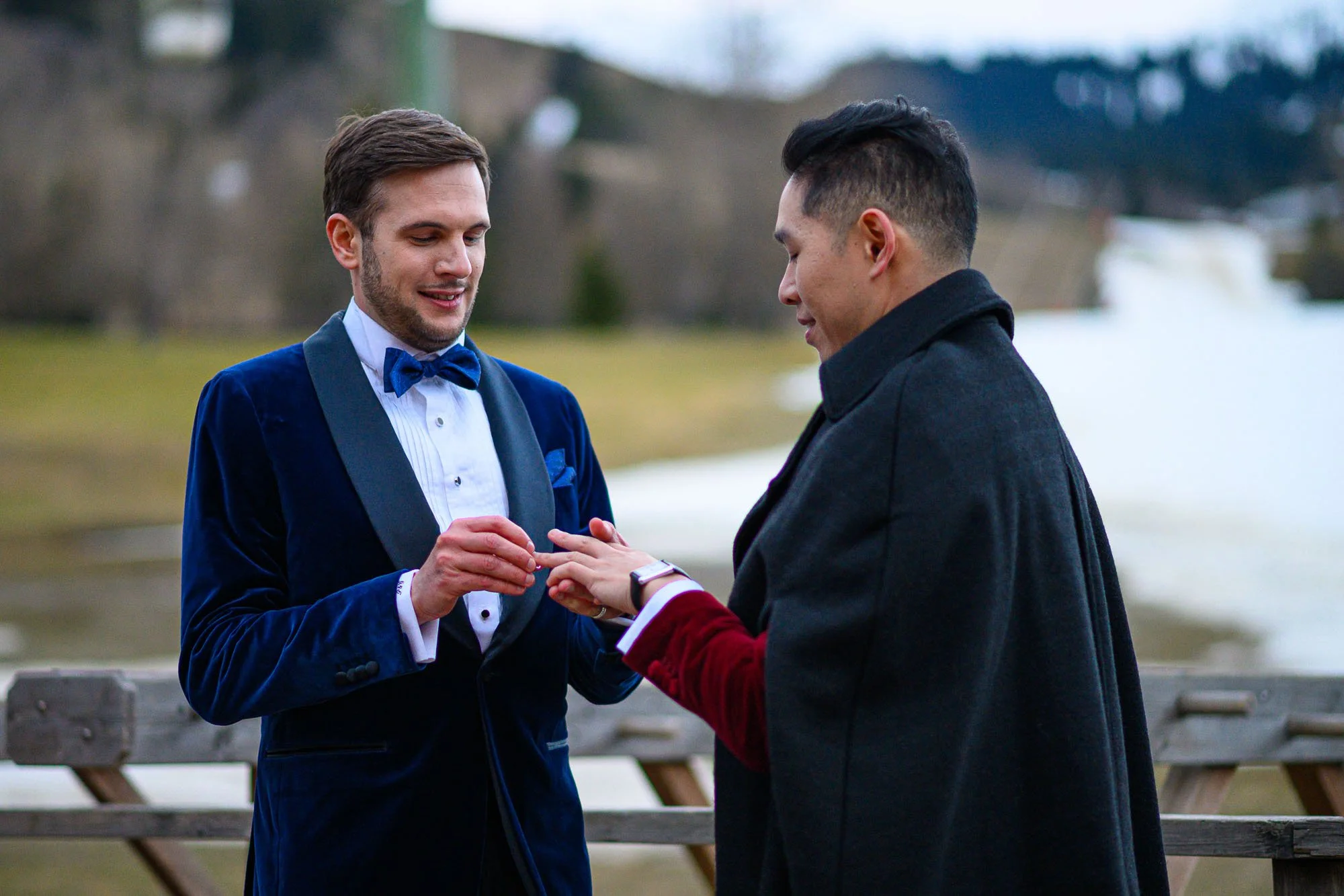 Deux hommes lors d'une cérémonie de mariage en plein air, échangeant des bagues, avec un plan d'eau et un paysage naturel en arrière-plan.