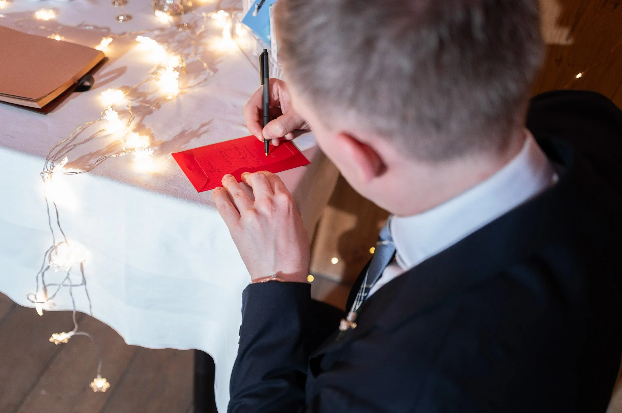 Un homme écrit une carte de vœux rouge à une table décorée avec des guirlandes lumineuses