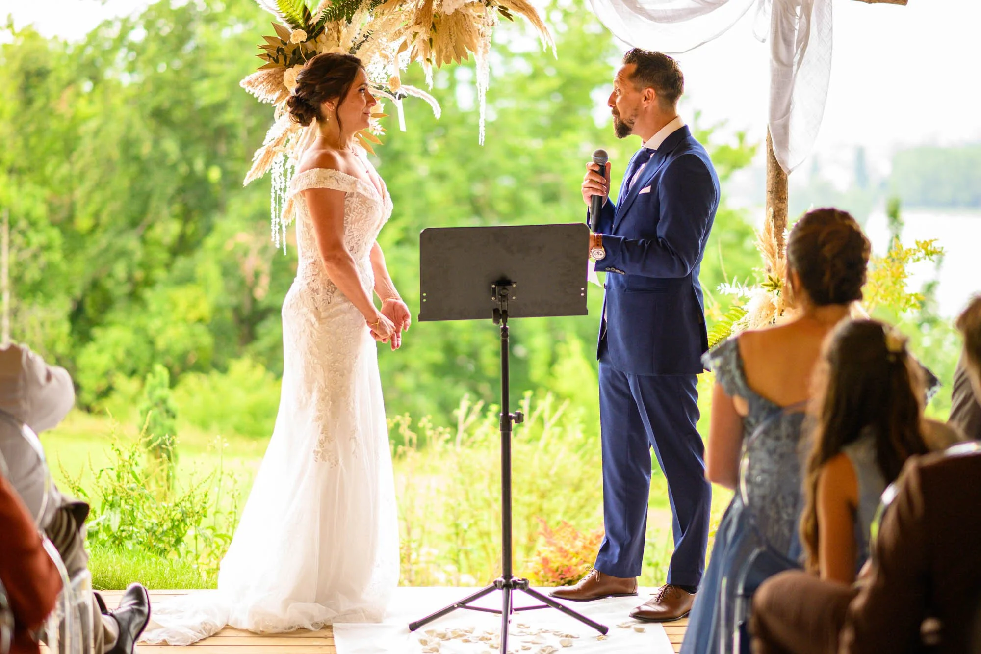 Un mariage en plein air avec une femme en robe de mariée et un homme en costume bleu échangeant leurs vœux sous une arche décorée de fleurs et de feuillage, entourés d'invités.
