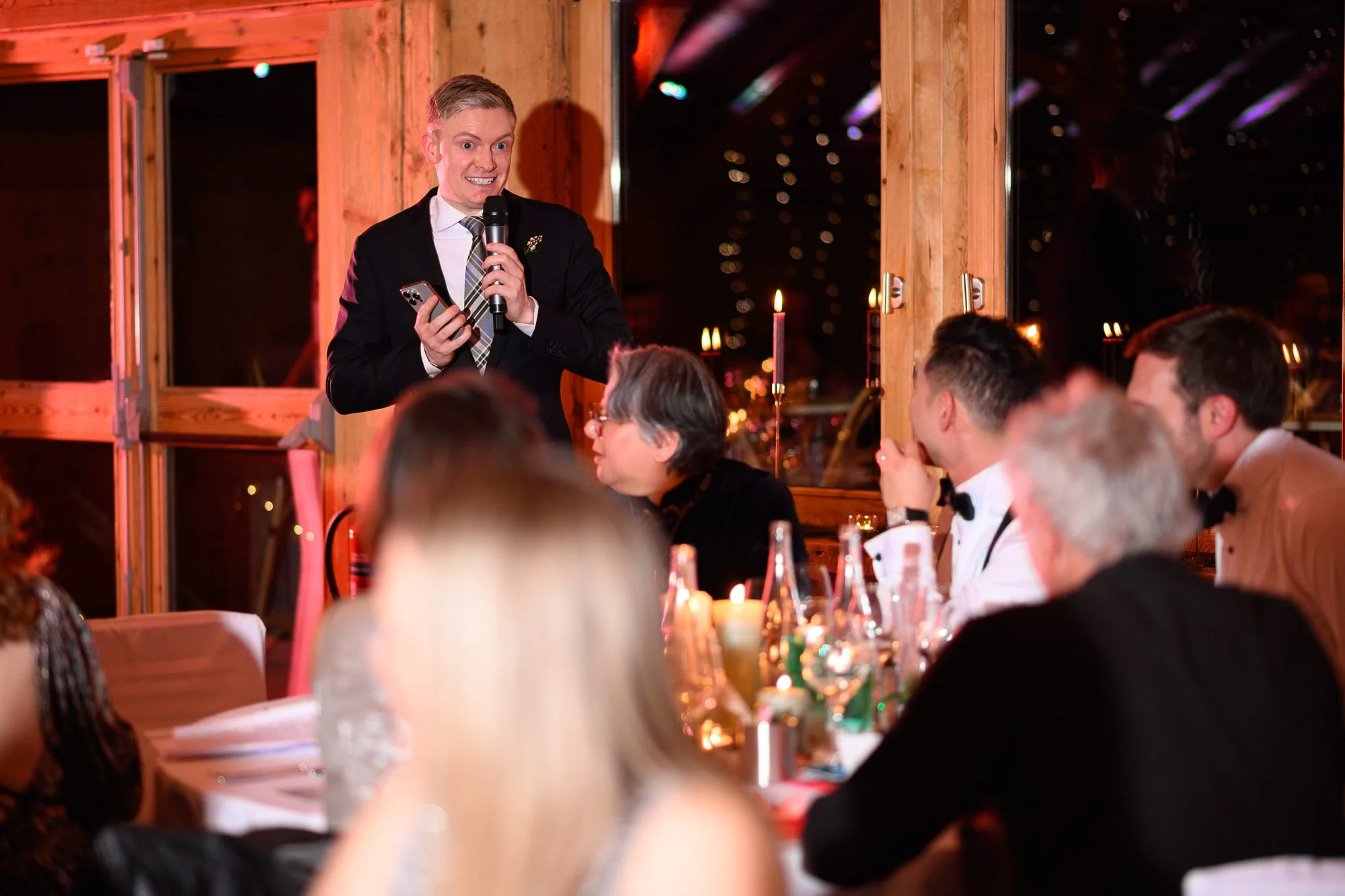 Un homme en costume noir parle dans un microphone lors d'un dîner formel, entouré de convives assis à une table avec bougies dans un décor en bois.