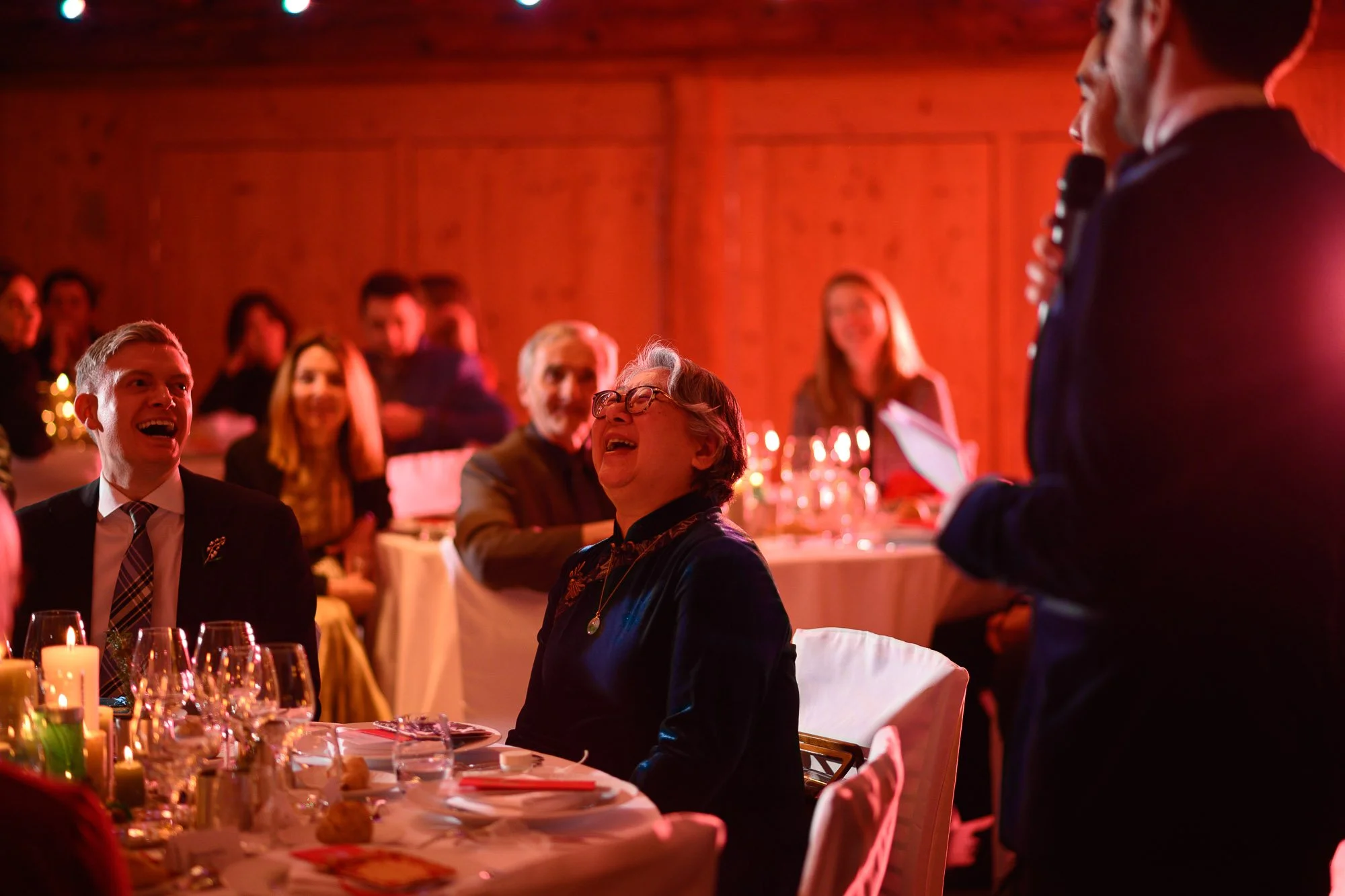 Groupe de personnes assises autour d'une table lors d'un dîner, riant et souriant, dans une ambiance chaleureuse avec éclairage tamisé.