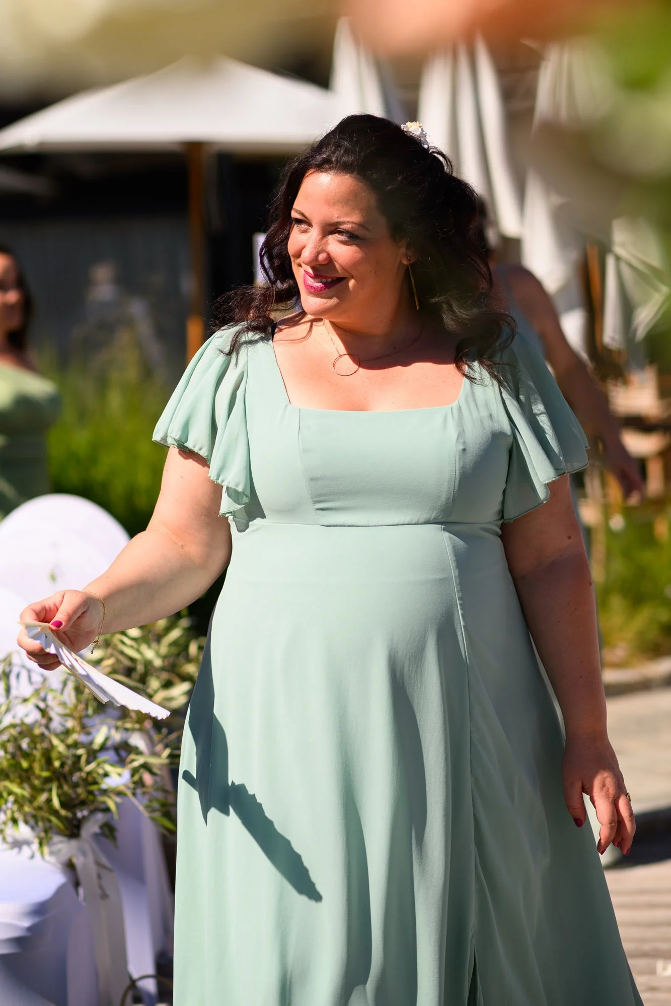 Femme souriante en robe verte claire à une cérémonie en extérieur, tenant un carnet ou une brochure, avec parasols en toile blanche en arrière-plan.