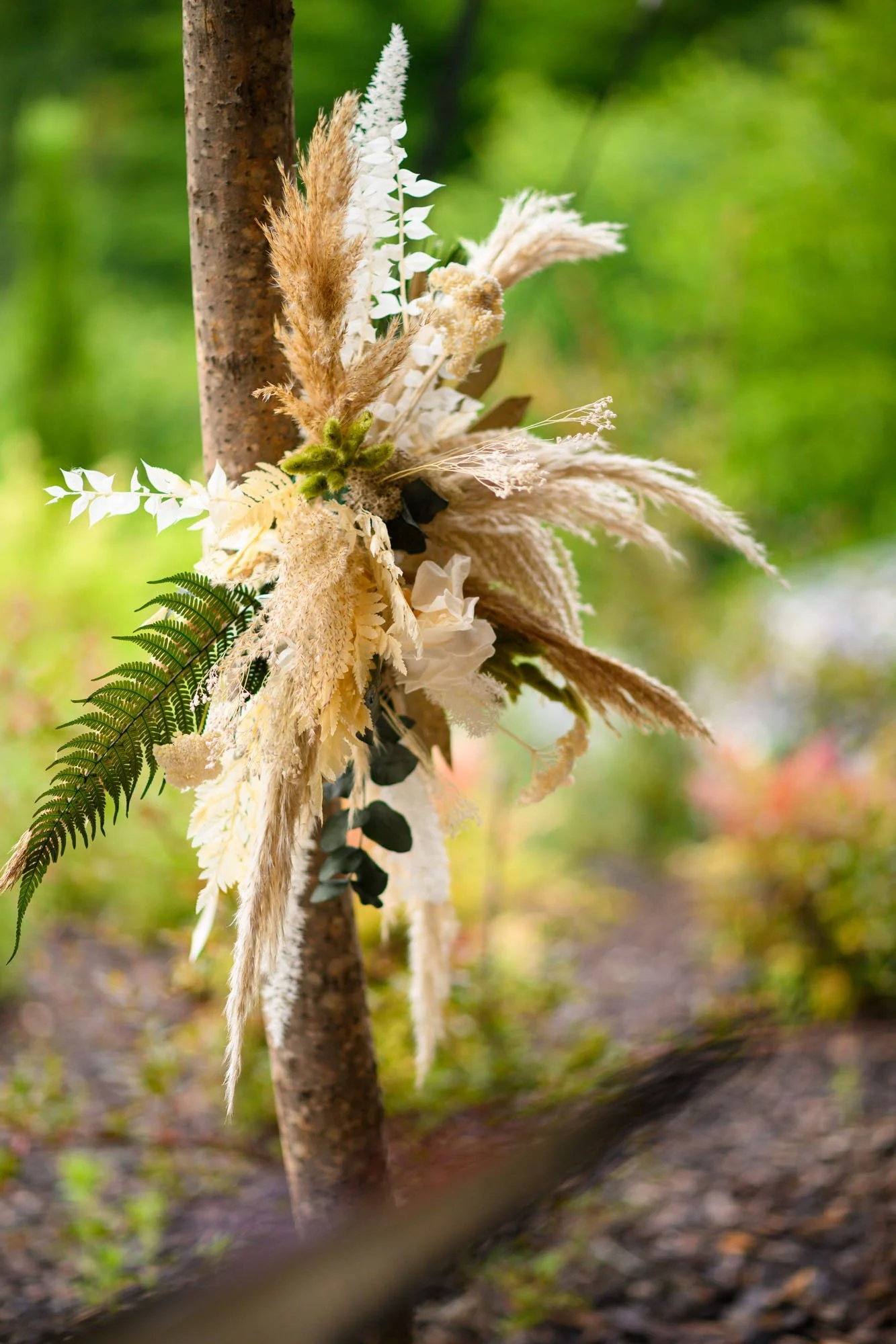 Arranjo floral suspendu à un arbre, comprenant des feuilles de fougère, des pampas blanches, brunes et beiges, avec un fond de forêt verte.