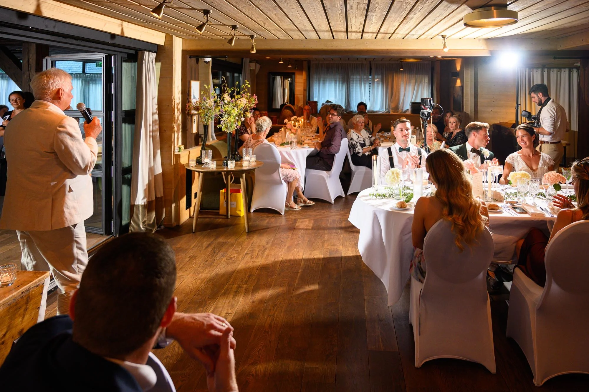 Un repas de mariage avec des invités assis autour d'une table, une personne parlant au micro, un décor chaleureux en bois, et un espace bien éclairé.