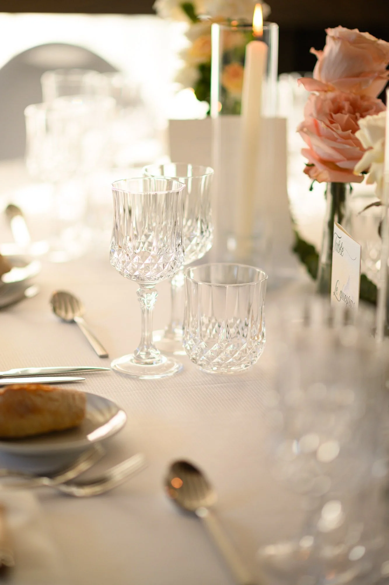 Table élégamment dressée avec des verres en cristal, une bougie, des fleurs roses et un menu, pour un événement ou un dîner formel.