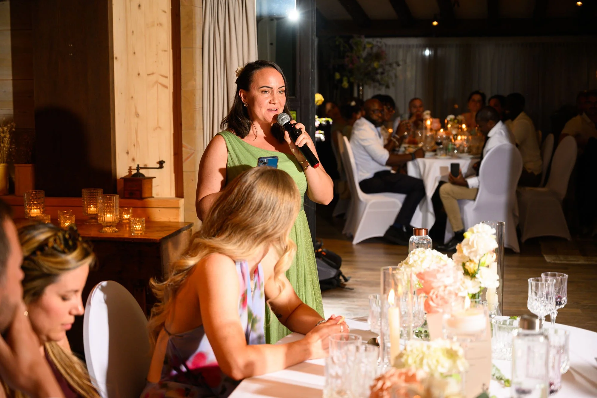 Une femme en robe verte parlant lors d'un événement, entourée de personnes assises à une table avec des décorations florales et des bougies, dans une salle éclairée chaleureusement.