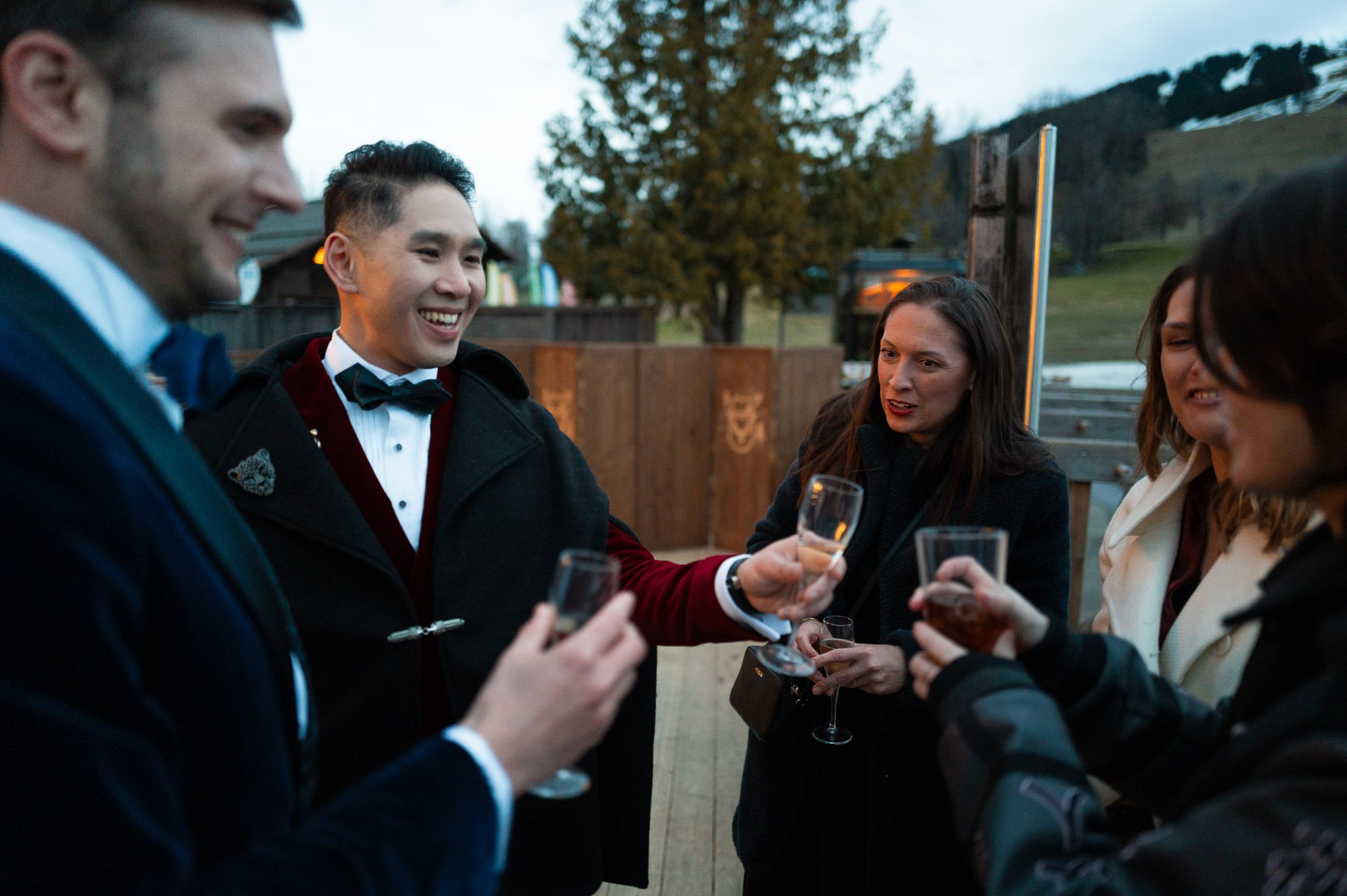 Groupe de cinq personnes lors d'une fête en plein air, portant des tenues elegantes, s'abreuvent et échangent des verres, avec un décor de nature et des montagnes en arrière-plan.