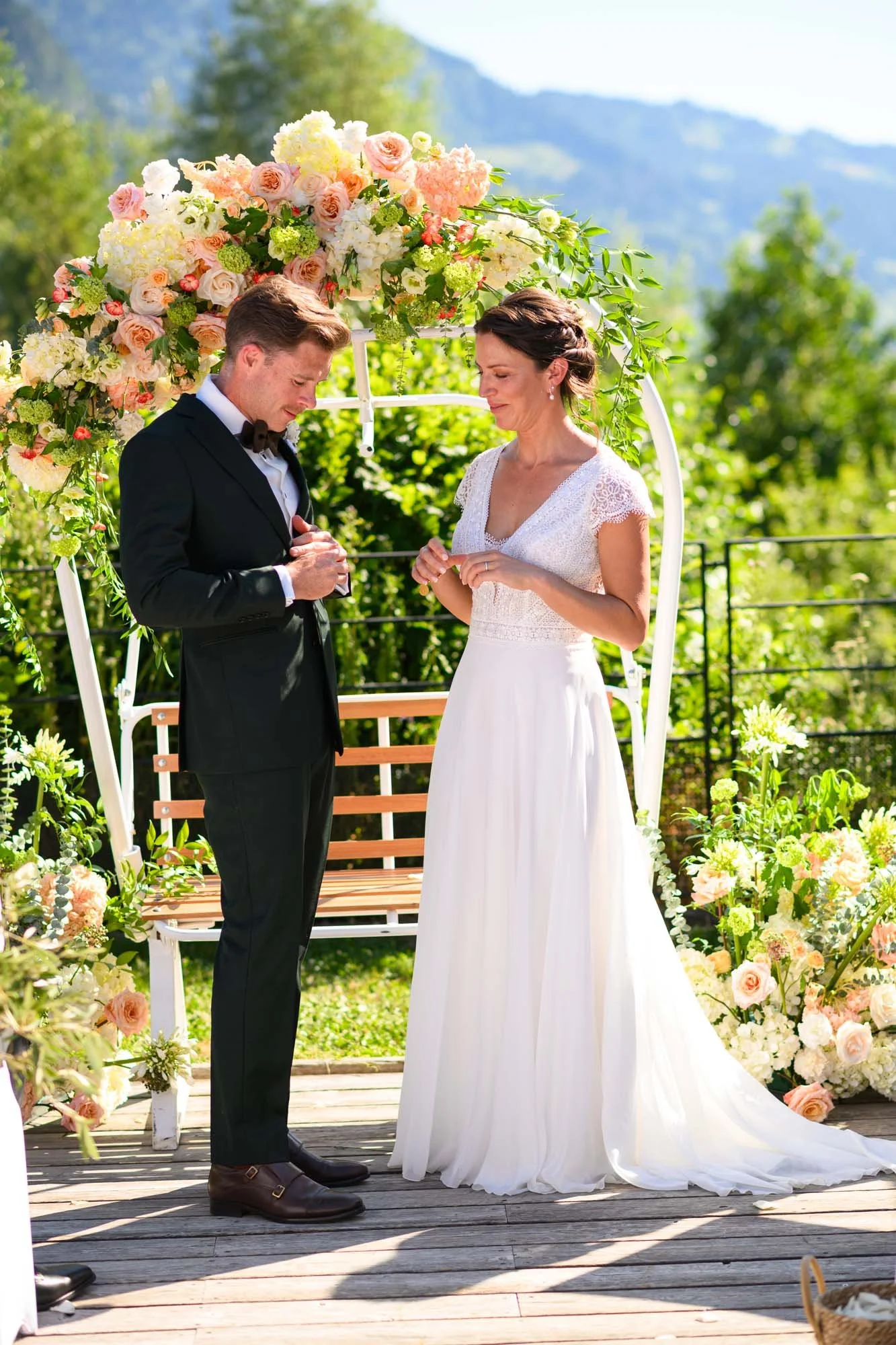 Un couple marié échangeant leurs vœux de mariage lors d'une cérémonie en plein air, sous un décor floral avec un siège en bois en arrière-plan, entourés de verdure et de montagnes en toile de fond.