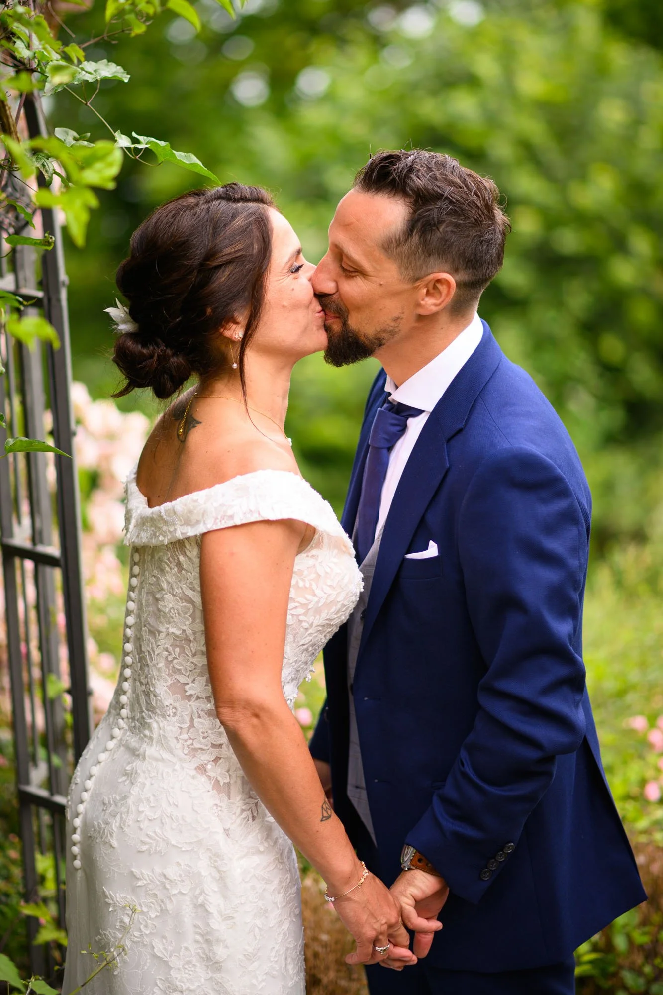 Un couple en costumes de mariage s'embrasse dans un jardin vert entouré de plantes et de fleurs.