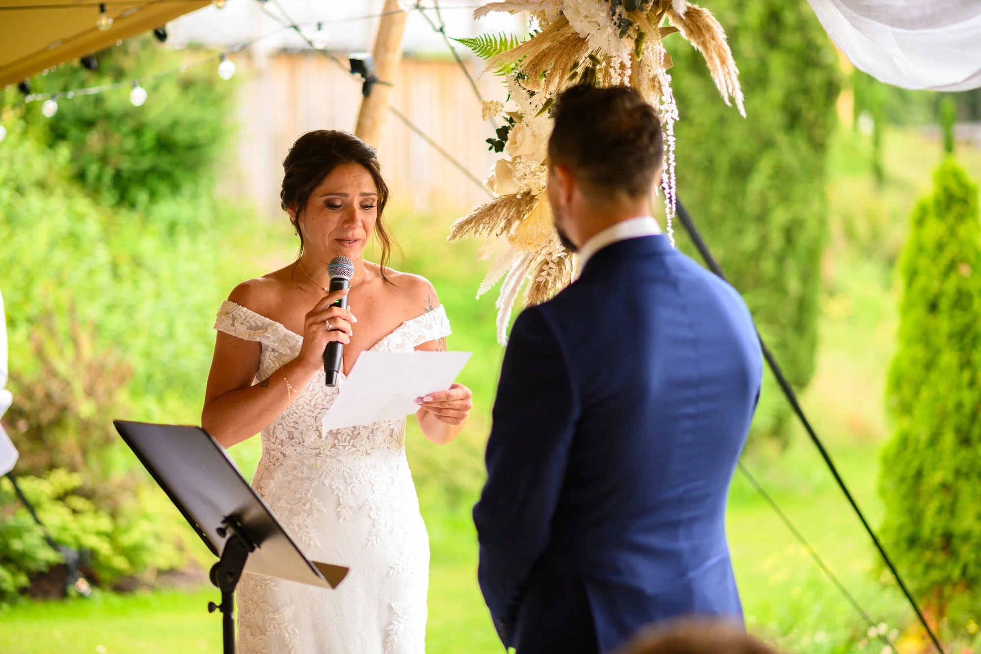 Une femme en robe de mariage lisant un vœu ou un discours, en face d'un homme en costume lors d'une cérémonie de mariage en plein air.