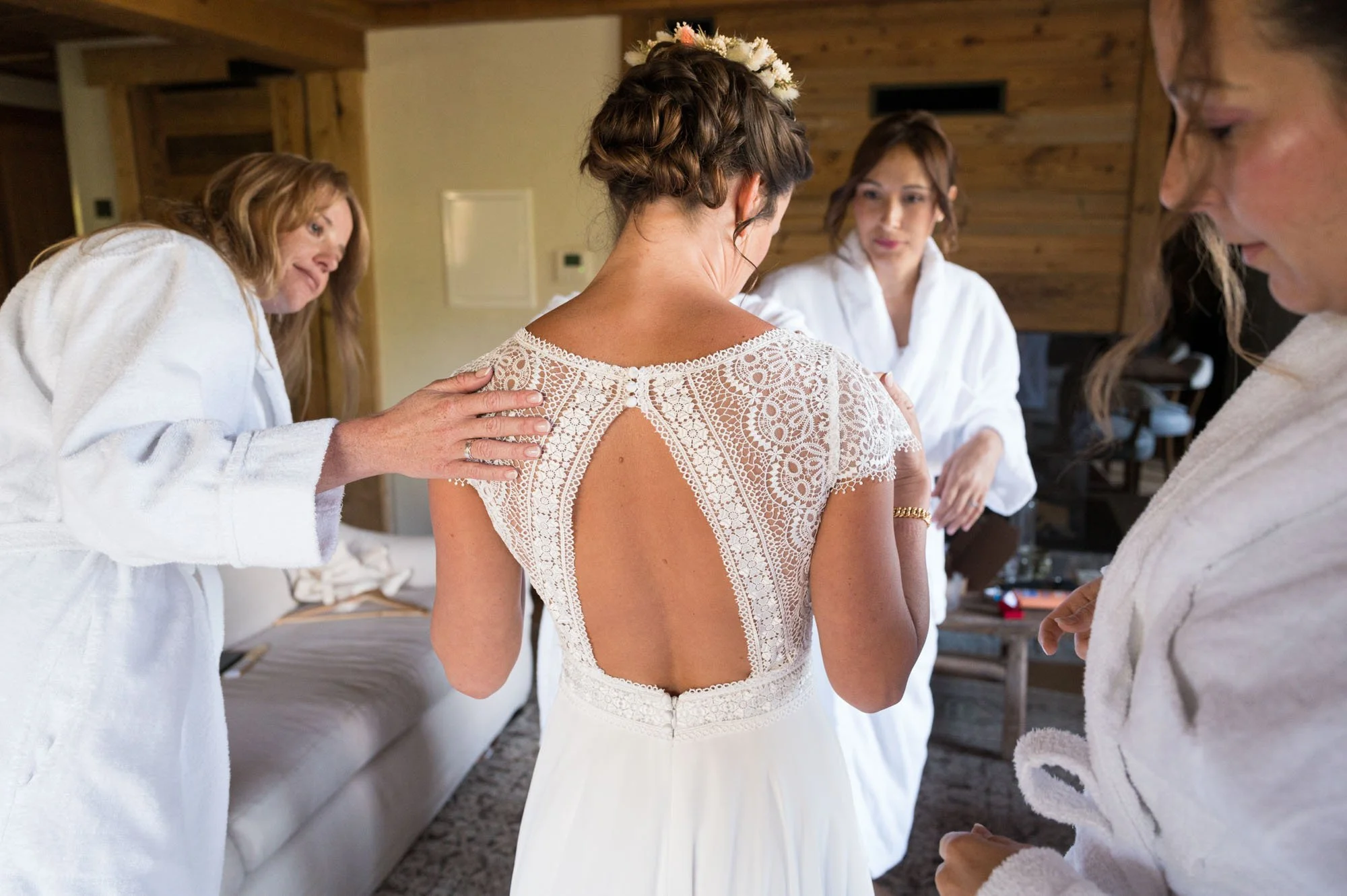 Une mariée en robe blanche avec un dos en dentelle, entourée de femmes en peignoirs blancs aidant à se préparer dans une pièce en bois.