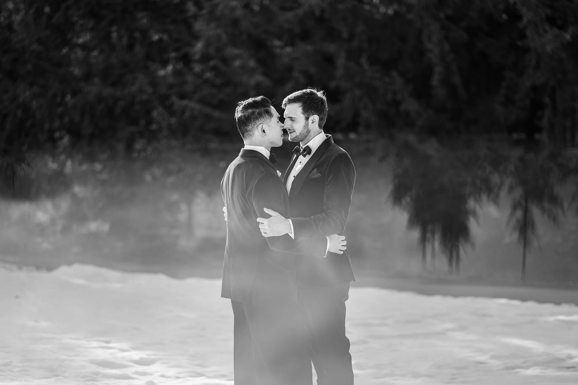 Deux hommes en costumes de mariage, en train de se tenir face à face et de se regarder tendrement, en extérieur. Black and white.