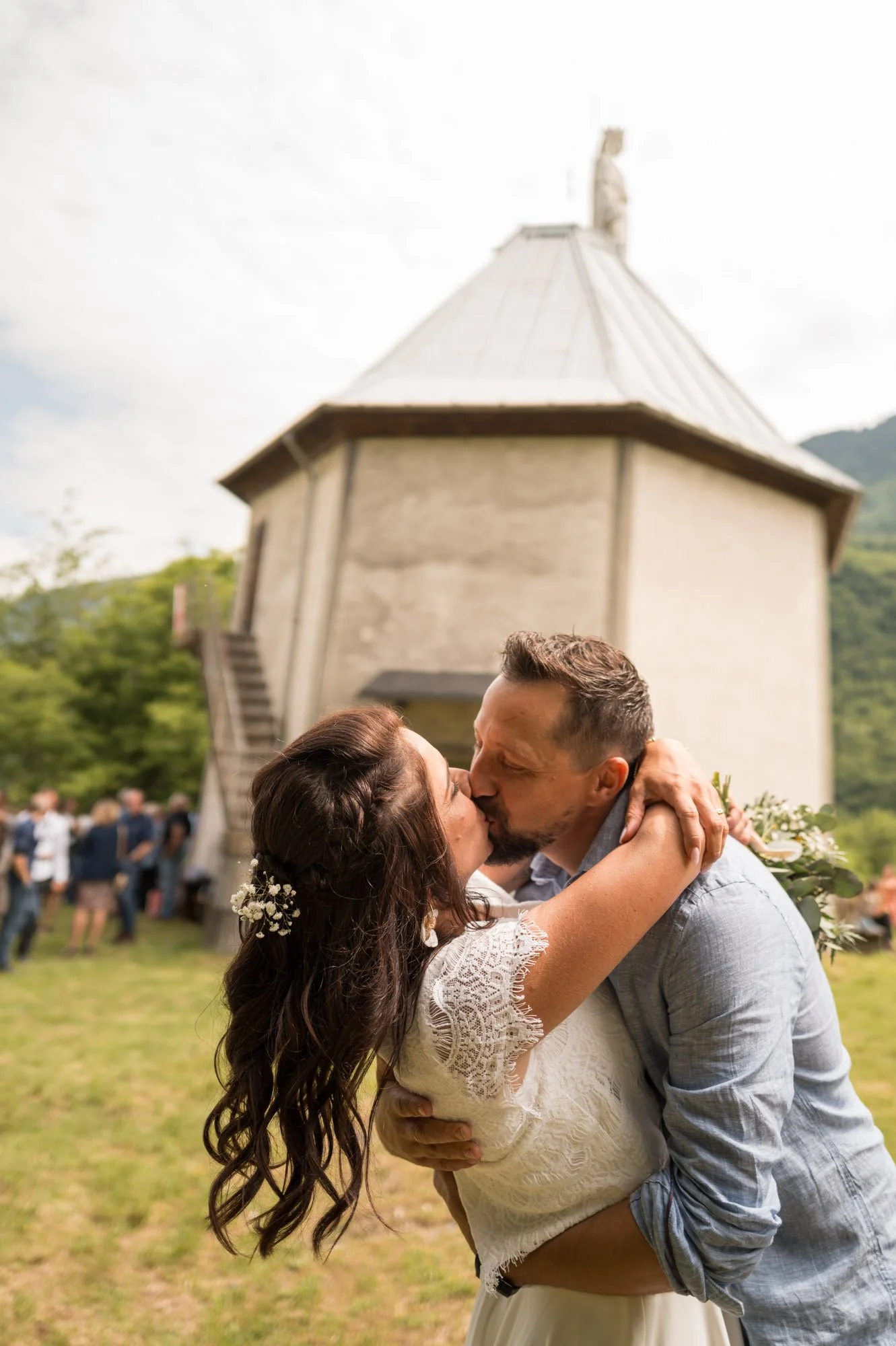 Un couple lors de leur mariage qui s'embrasse, avec une église en pierre en arrière-plan et des invités en arrière-plan.