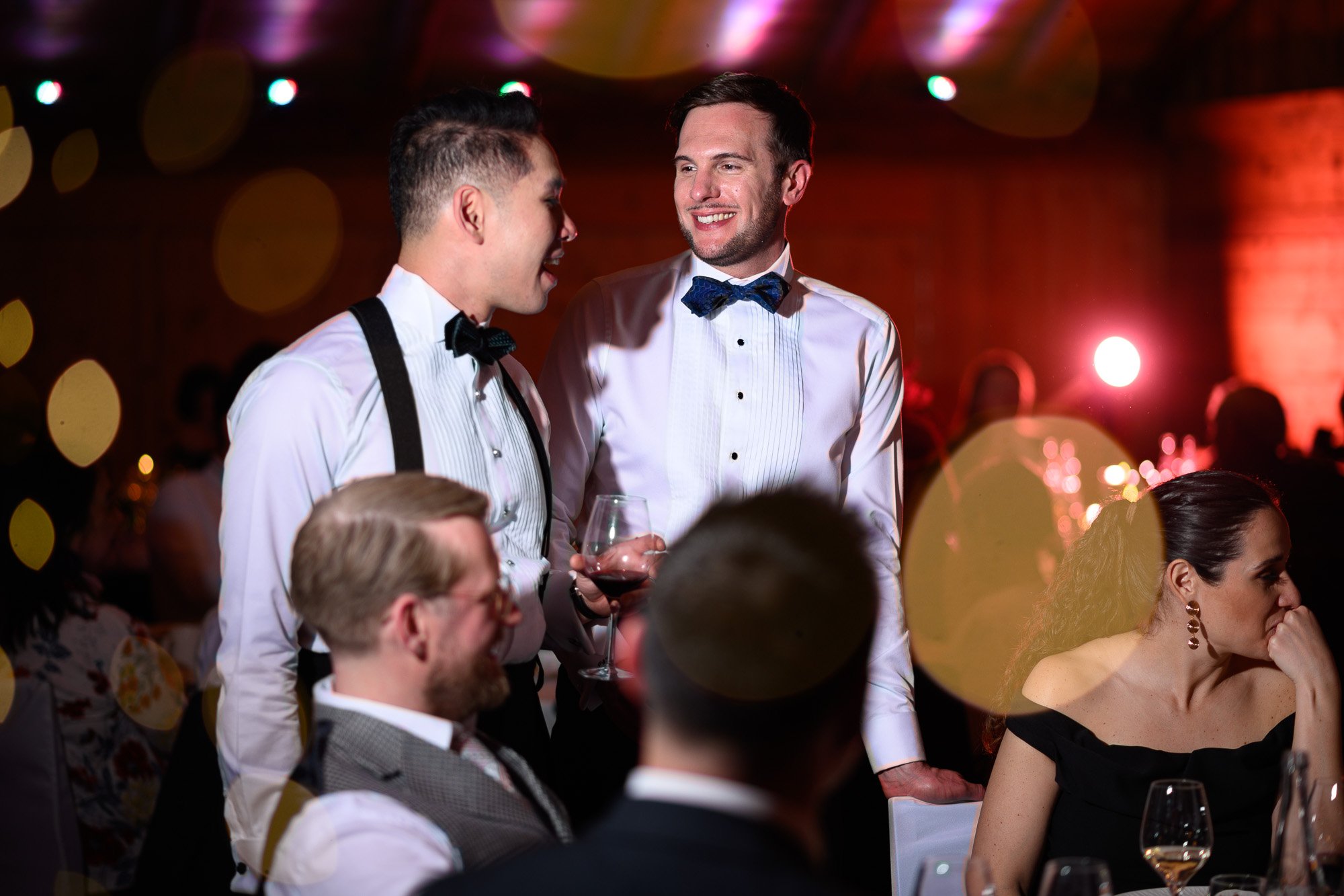 Deux hommes en tenues de soirée discutent et rient lors d'un événement, entourés de personnes assises à une table, avec un fond coloré et éclairage tamisé.