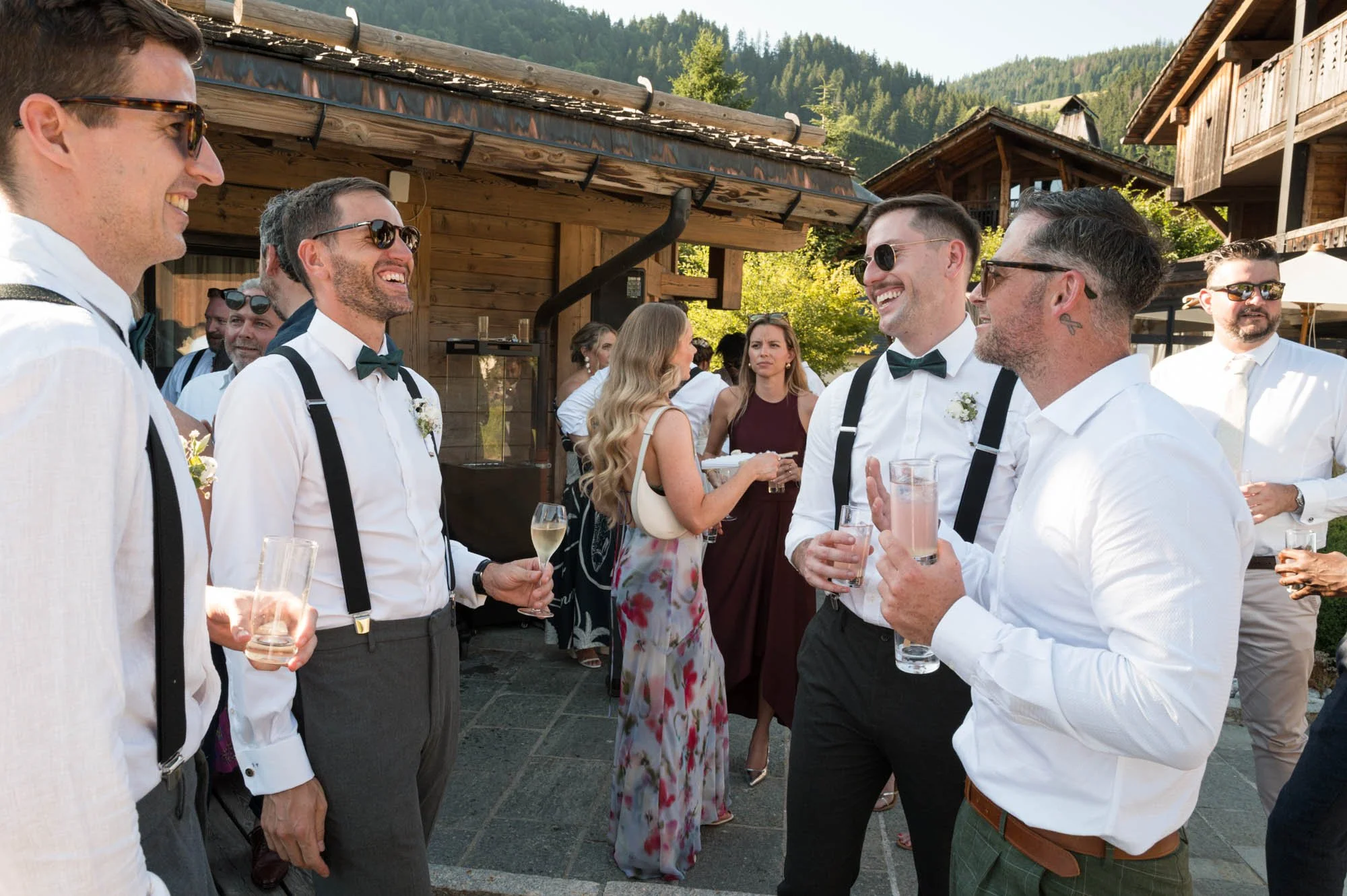 Groupe de personnes habillées élégamment, célébrant et discutant lors d'une fête en extérieur, avec des bâtiments en bois et des montagnes en arrière-plan.