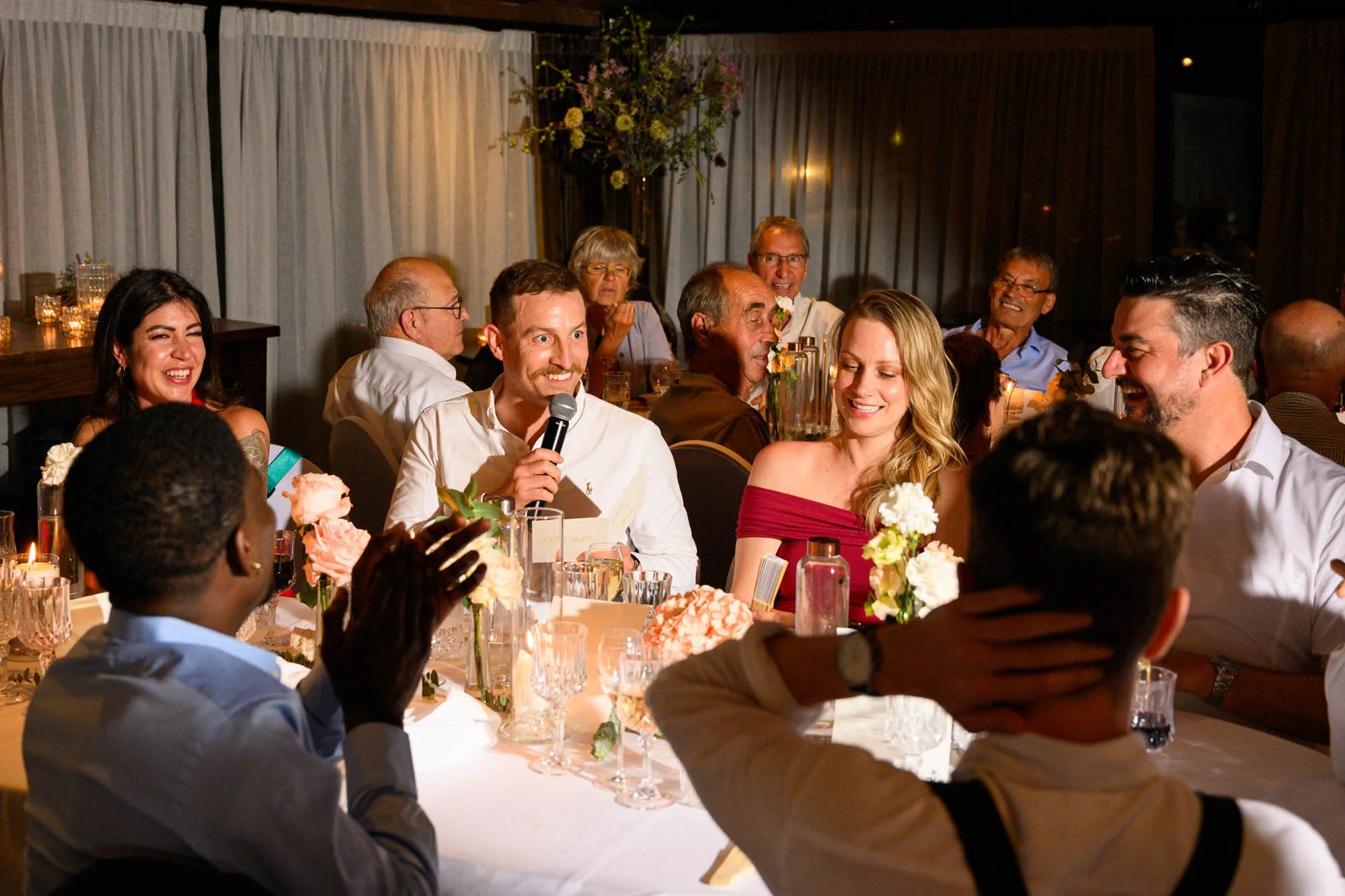 Groupe de personnes riant et discutant autour d'une table lors d'une célébration ou d'un dîner, décorée de fleurs et de bougies.