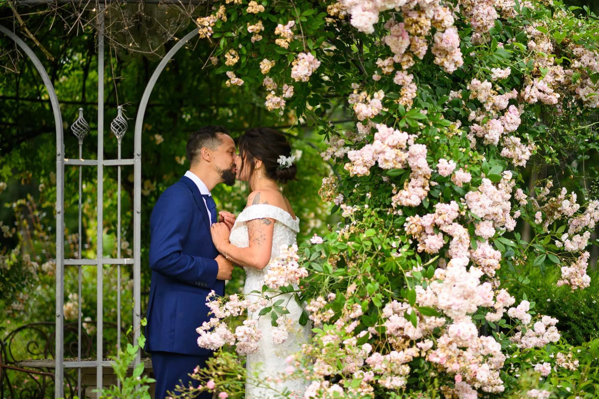 Un couple de mariés en costume et en robe de mariée s'embrassent dans un jardin entouré de fleurs roses et de verdure.