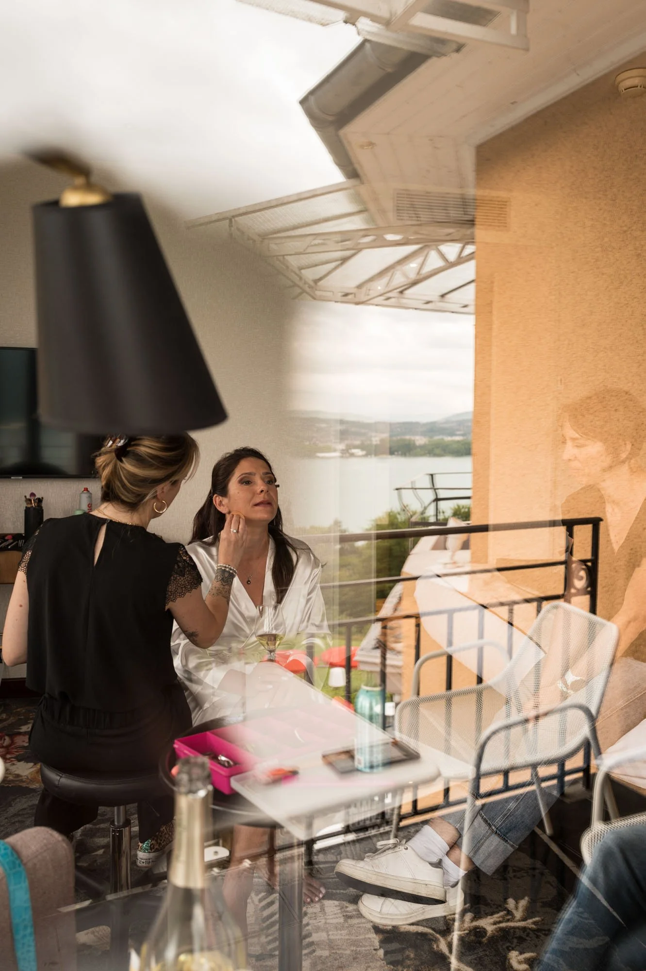 Une femme fait un maquillage à une autre femme qui est assise à une table avec un verre de vin blanc, avec un décor de balcon et une vue sur la nature en arrière-plan.