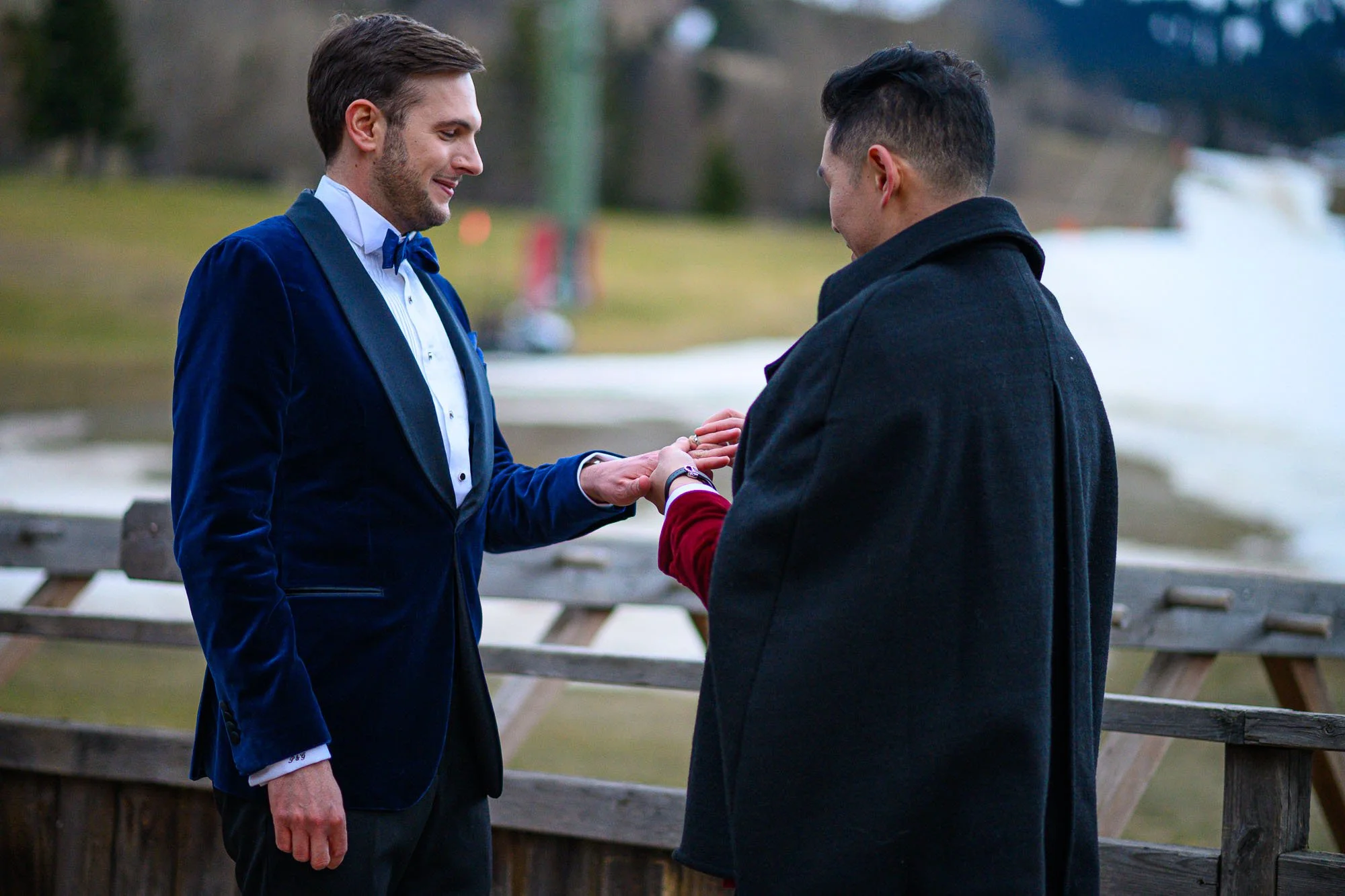 Deux hommes, l'un en costume bleu avec une chemise blanche et une cravate, l'autre en manteau noir, échangent des alliances face à un lac, lors d'une cérémonie de mariage.