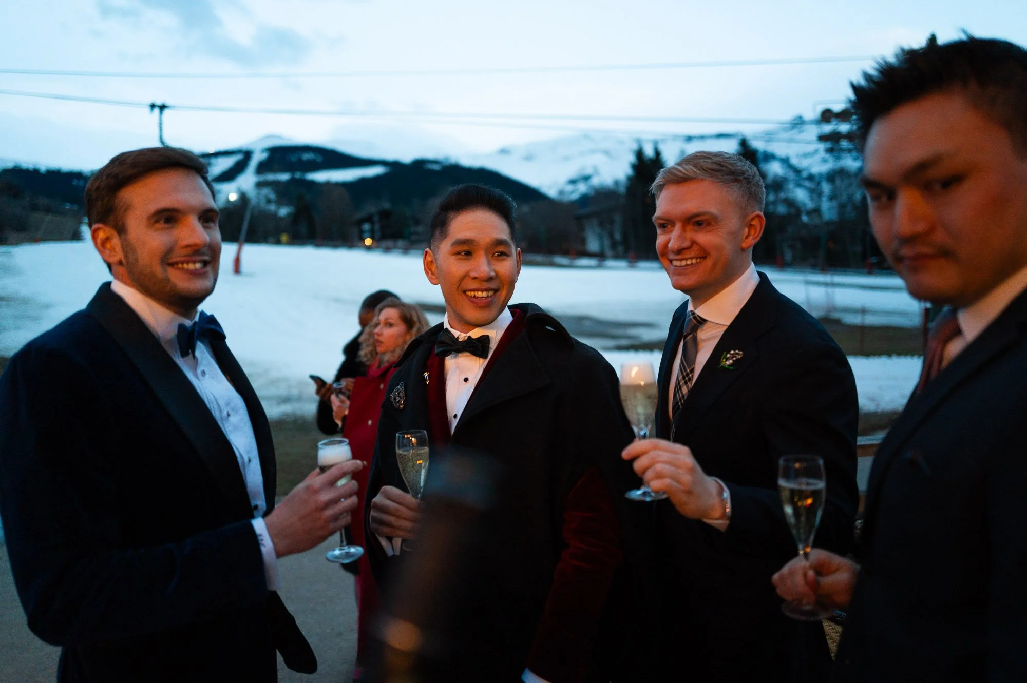 Groupe d'hommes en costume lors d'une fête en plein air avec des montagnes enneigées en arrière-plan.