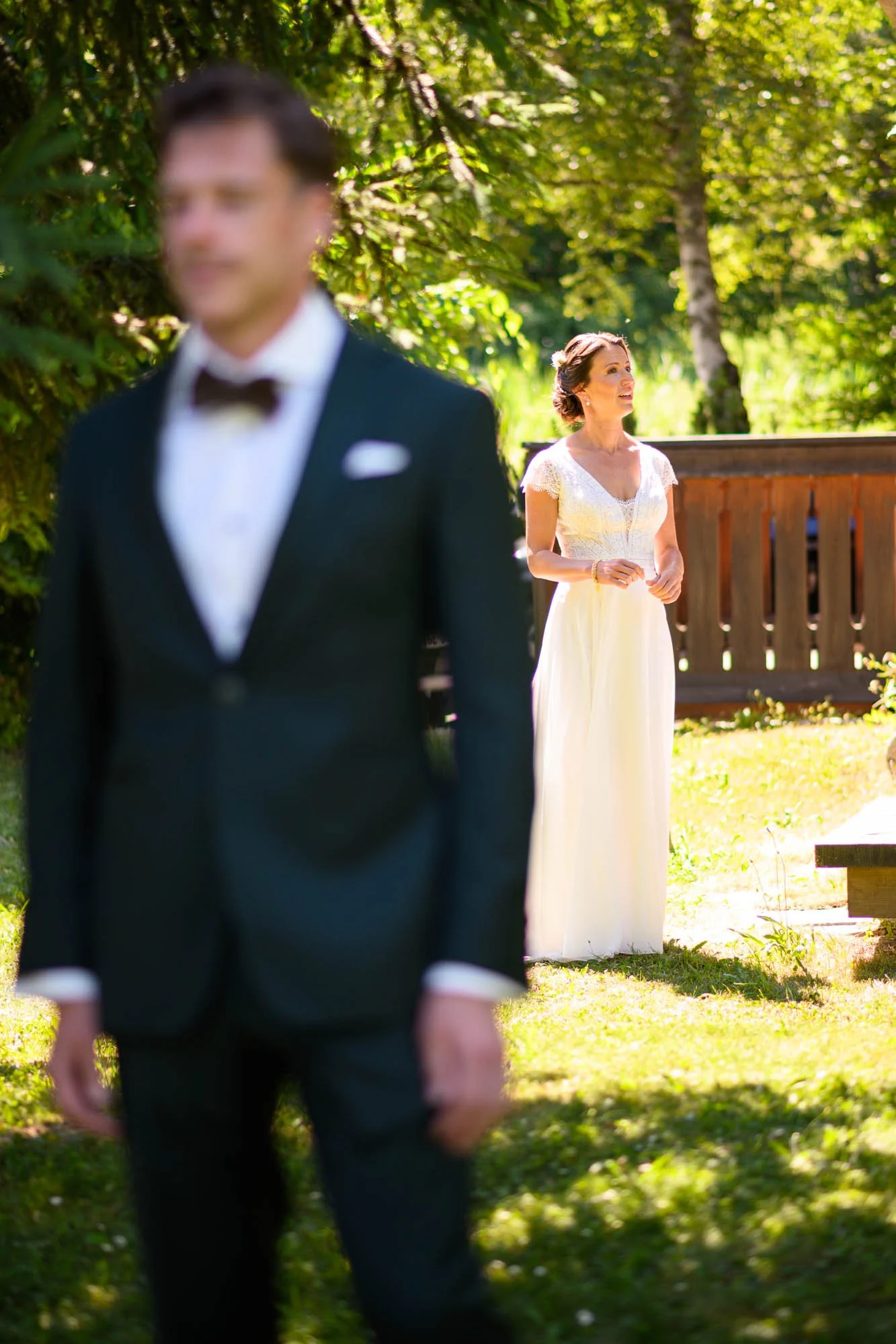 Un homme en costume de mariage et une femme en robe blanche debout dans un jardin ensoleillé, avec des arbres en arrière-plan.
