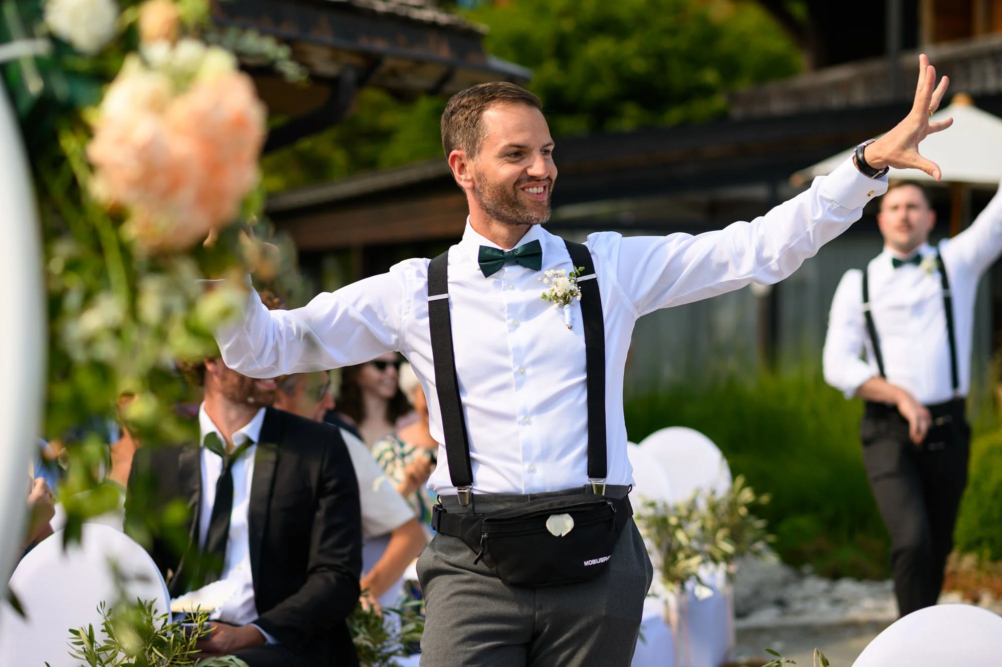 Un homme souriant en costume blanc et nœud papillon vert lors d'un mariage en plein air, avec d'autres personnes habillées formellement en arrière-plan.